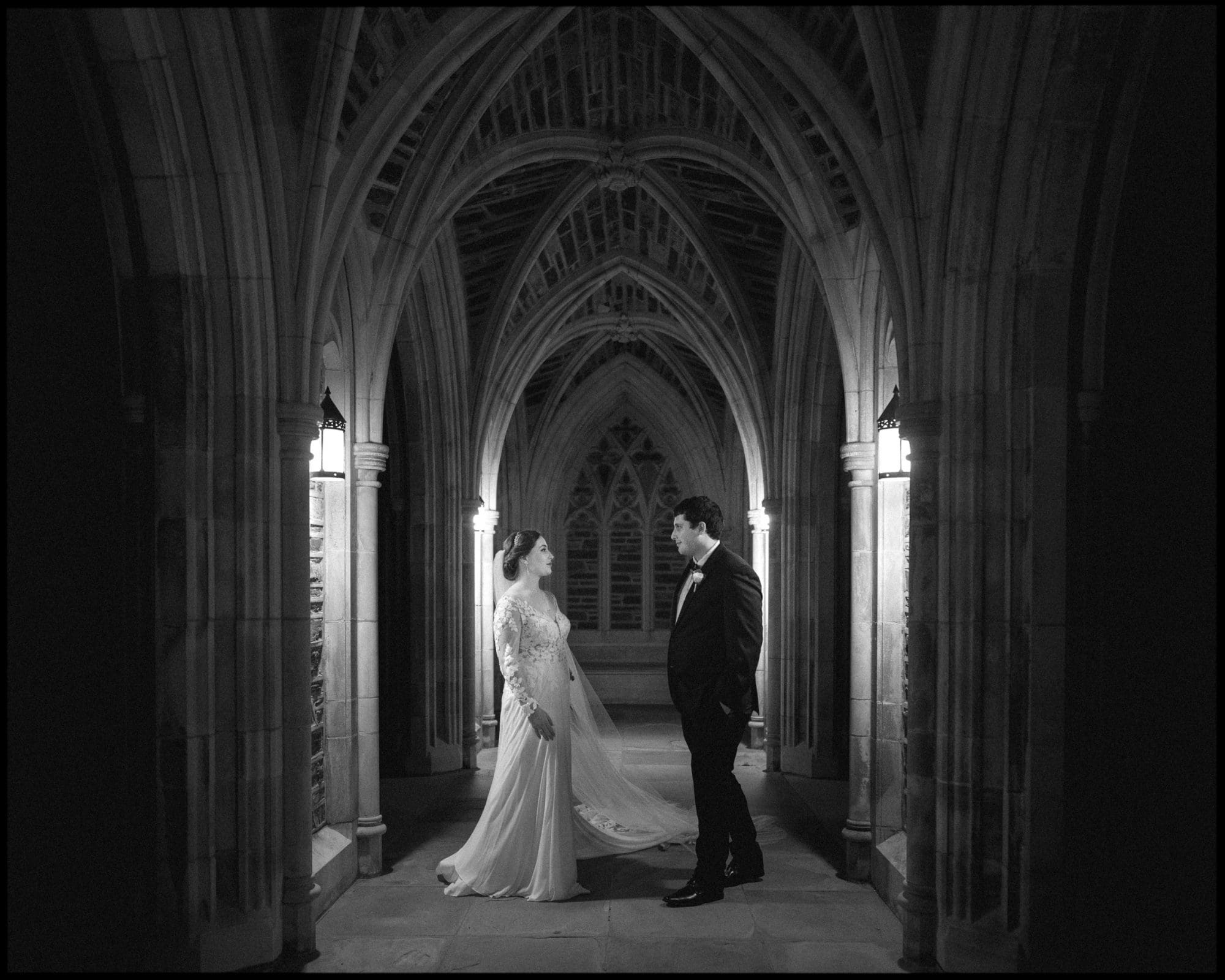 A bride and groom stand facing each other in a dimly lit, arched corridor of Duke Chapel, surrounded by ornate stonework. The bride wears a flowing gown, the groom in a suit. Captured in black and white, this Duke Chapel wedding exudes a timeless and romantic atmosphere.