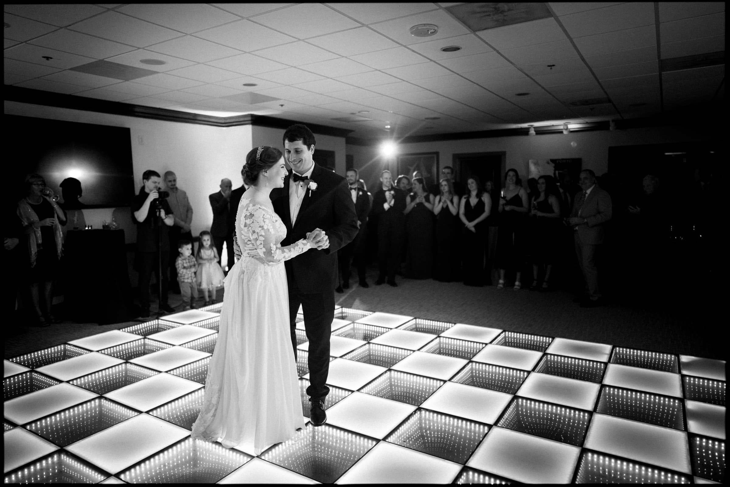 A bride and groom share their first dance on an illuminated checkered floor at the Duke Chapel wedding. They are surrounded by guests who watch and take photos, capturing the enchanting moment. The room is dimly lit, creating a romantic atmosphere that perfectly complements the occasion.