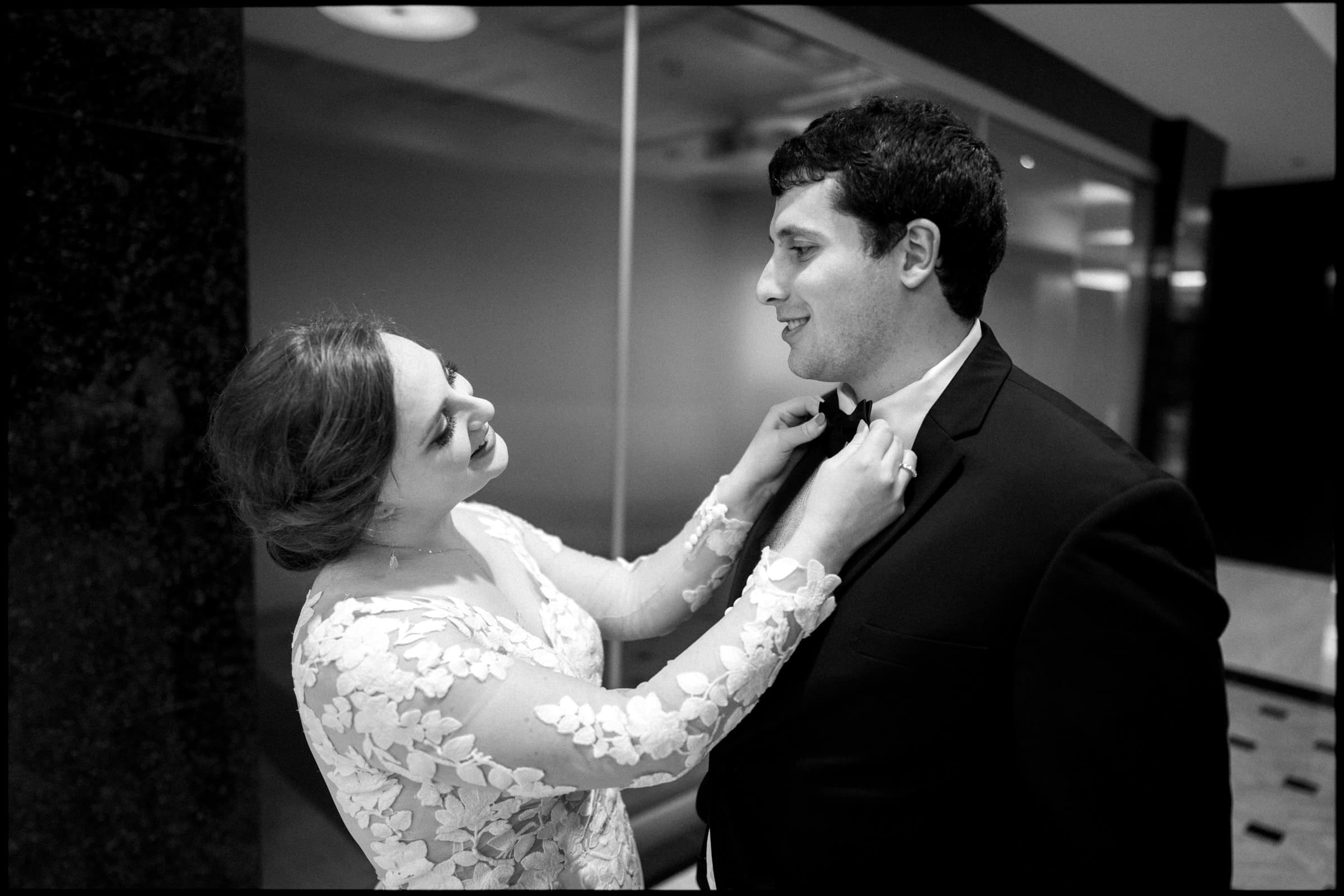 In an elegant indoor setting reminiscent of a Duke Chapel wedding, a bride in a lace dress adjusts the groom's bow tie. They exchange warm smiles, with him in a dark suit. Reflective surfaces and soft lighting add to the enchanting atmosphere.