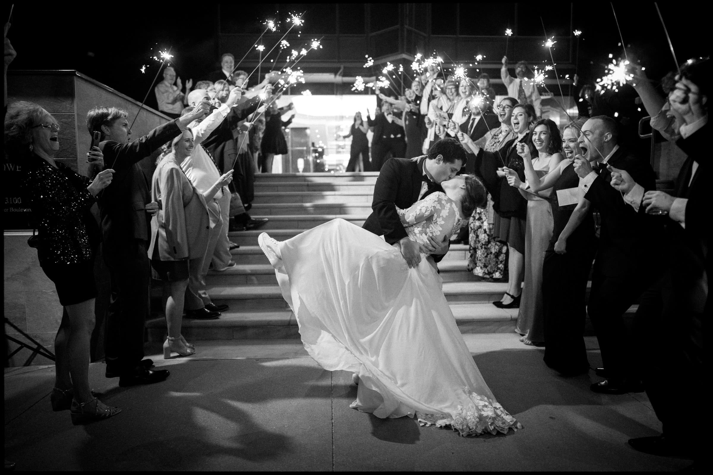 Amidst a joyful atmosphere, a bride and groom share a romantic kiss as the groom dips the bride on the grand staircase of Duke Chapel. Surrounded by cheering guests holding sparklers, they celebrate their enchanting nighttime wedding.