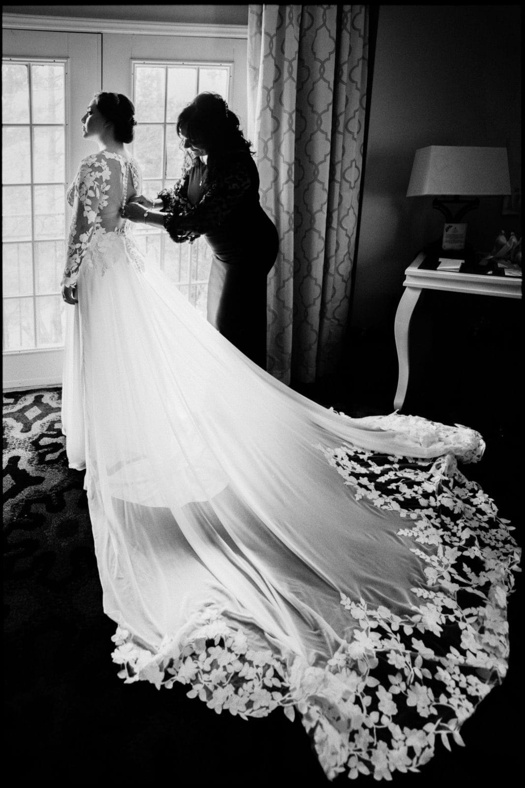A black and white photo captures a bride at a Duke Chapel wedding in a long, flowing gown with lace details, standing by a window. Another woman stands behind her, adjusting the dress. A small table and lamp are visible in the background.