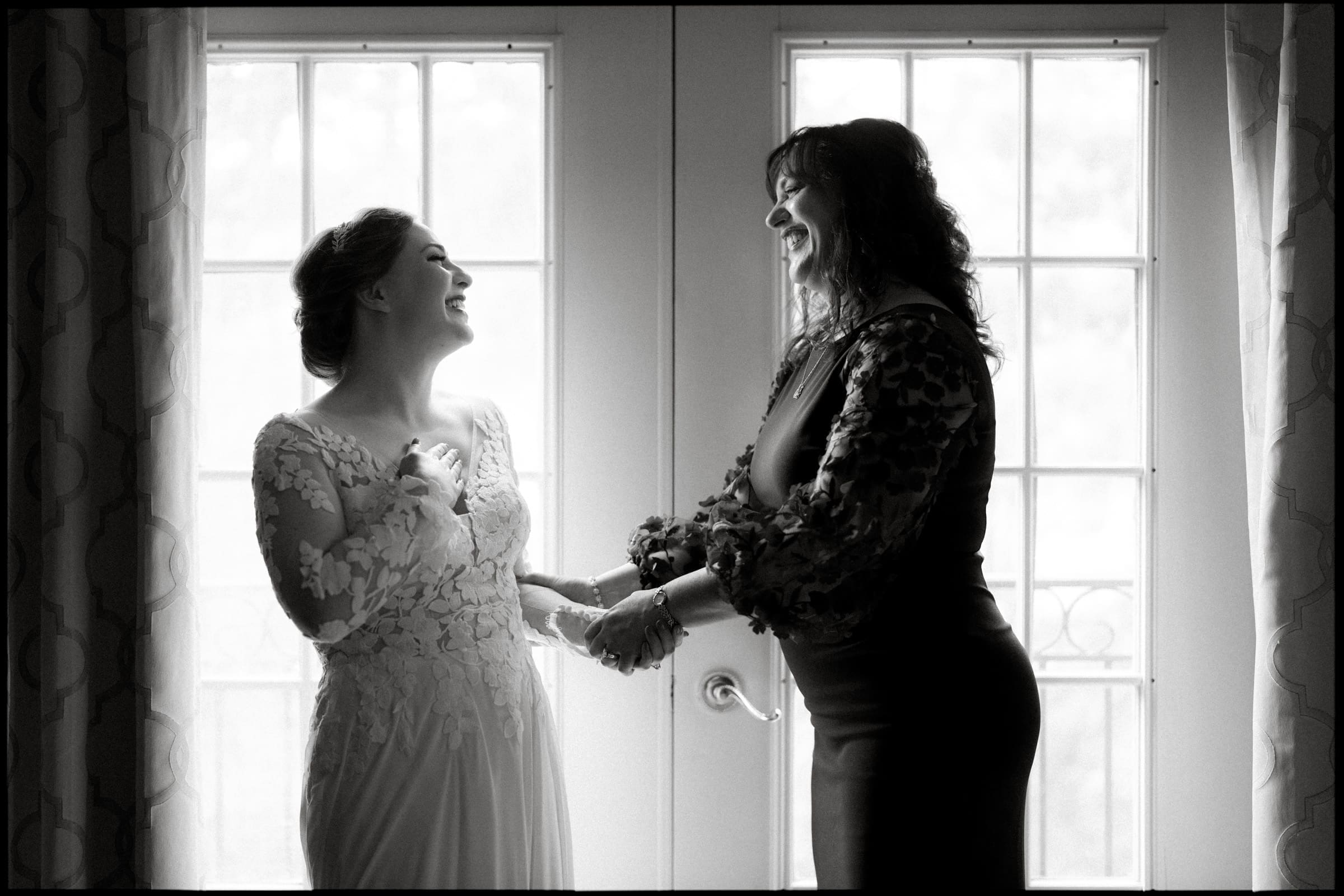 In the serene setting of Duke Chapel, a bride in a lacy gown shares a joyful moment with a woman in a dark dress. They stand before the bright window, smiling and gazing at each other lovingly.