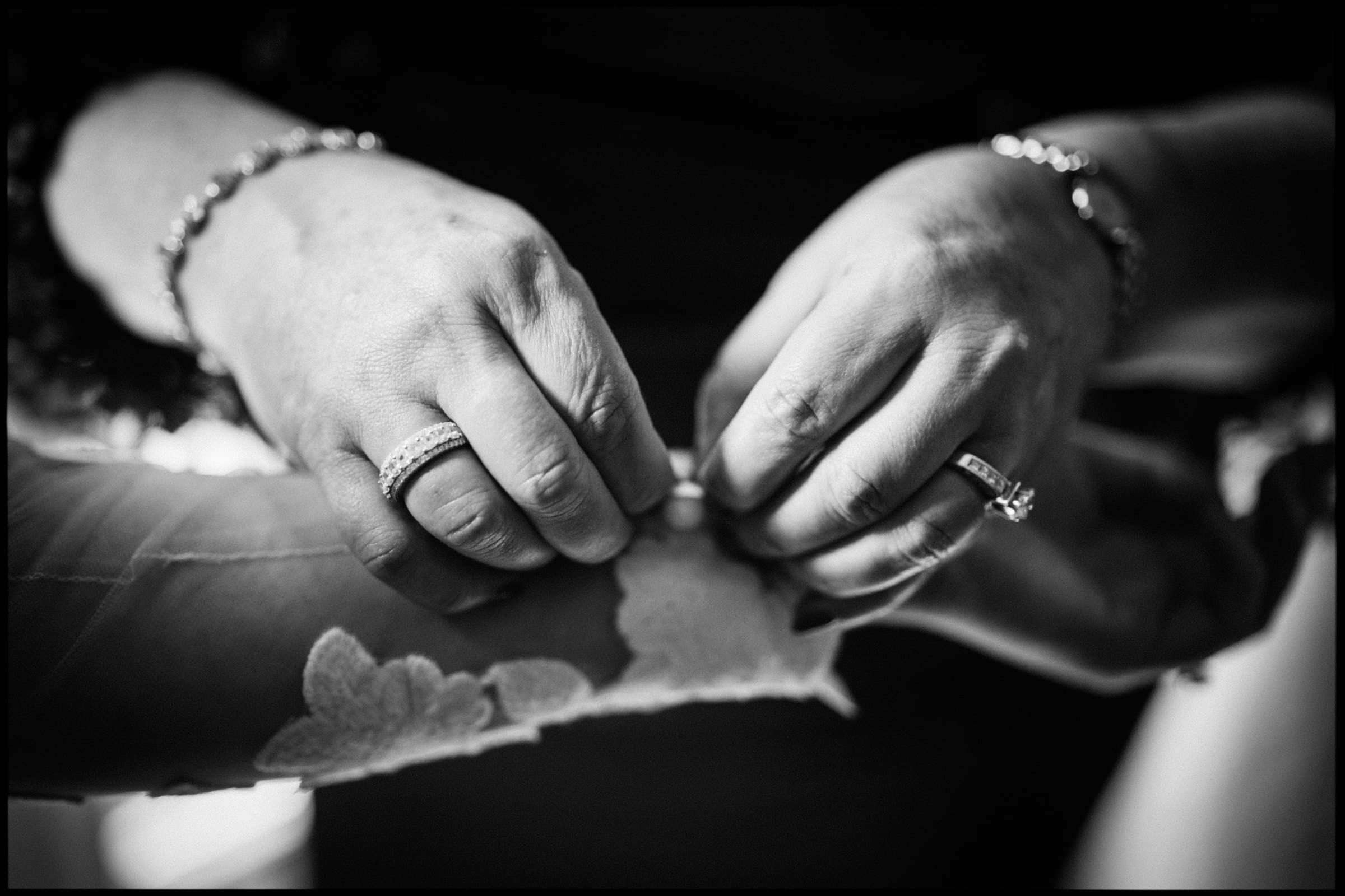 A close-up captures hands delicately fastening buttons on a lace sleeve, evoking the grace of a Duke Chapel wedding. The person wears rings and a bracelet, adding elegance to the black and white image.