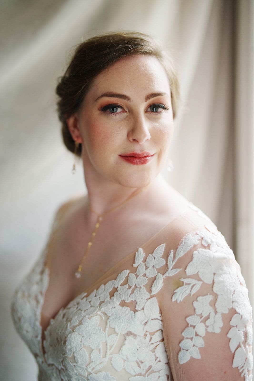 A bride in a white lace dress poses indoors at Duke Chapel, looking at the camera. Her hair is styled up, and she wears subtle makeup, drop earrings, and a delicate necklace. The background is softly blurred.