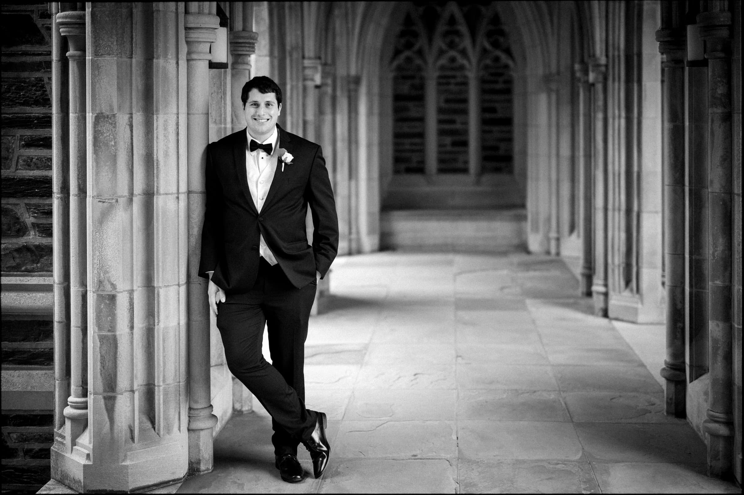 A man in a suit with a bow tie leans casually against a stone pillar in the Gothic-style elegance of Duke Chapel. The setting boasts ornate archways and detailed stonework, creating an atmosphere that's both formal and relaxed, perfect for capturing the essence of a timeless wedding.