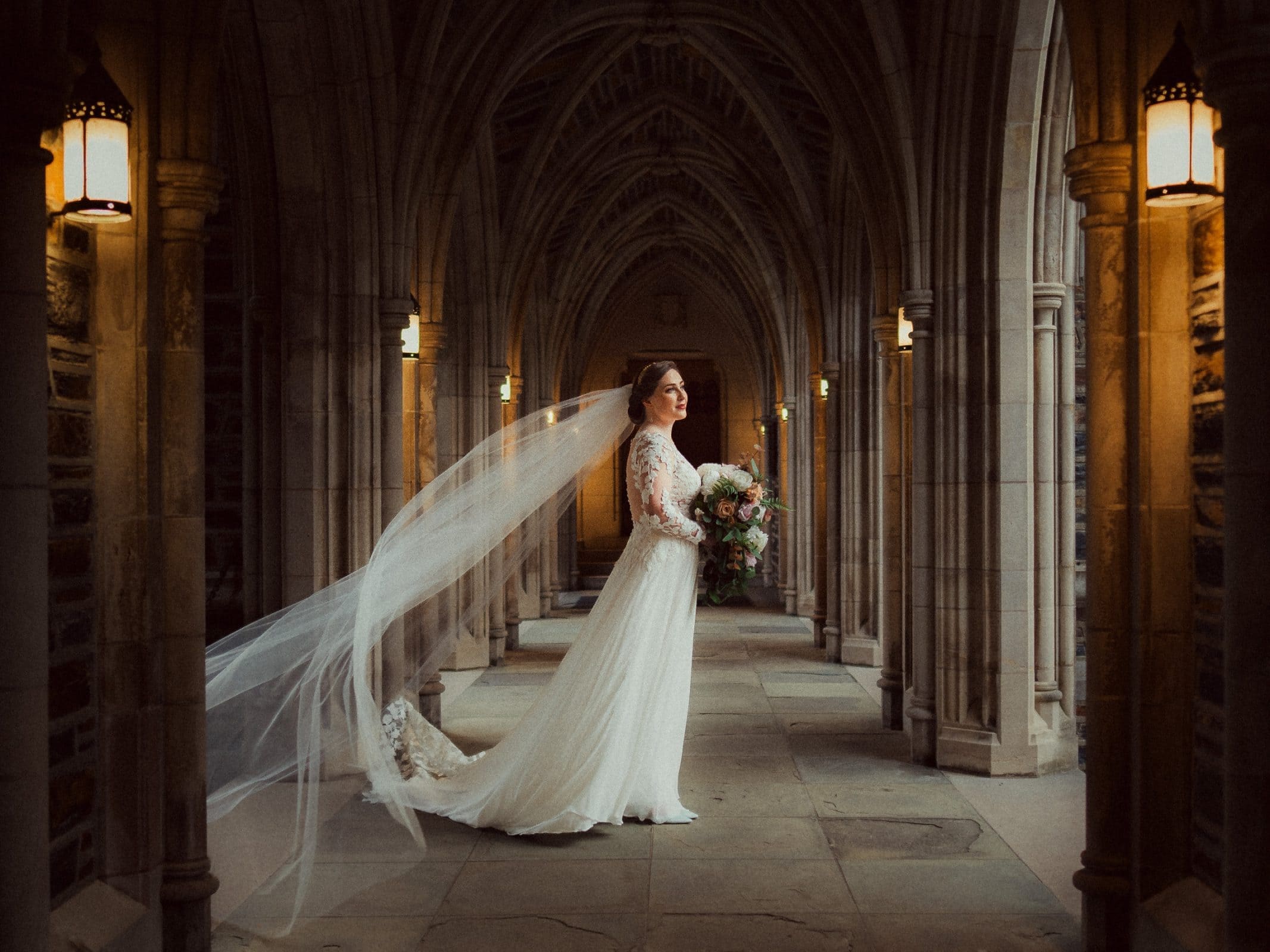 A bride in a long lace gown stands in the stone corridor of Duke Chapel, with arched ceilings and her bouquet in hand. Her veil flows elegantly behind her as warm lanterns light the romantic wedding setting.