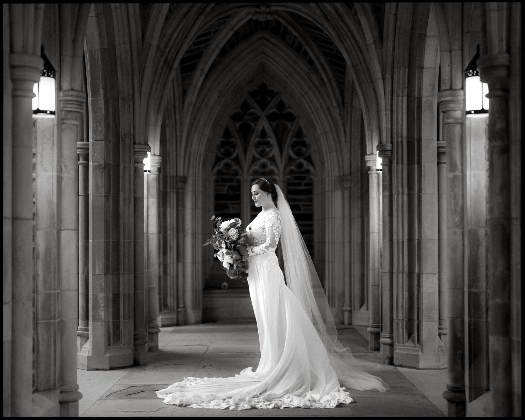 A bride in an elegant gown and veil stands holding a bouquet in a cathedral-like stone corridor, reminiscent of a Duke Chapel wedding. The scene is illuminated by soft lights, highlighting the arches and intricate architectural details. The mood is serene and timeless.