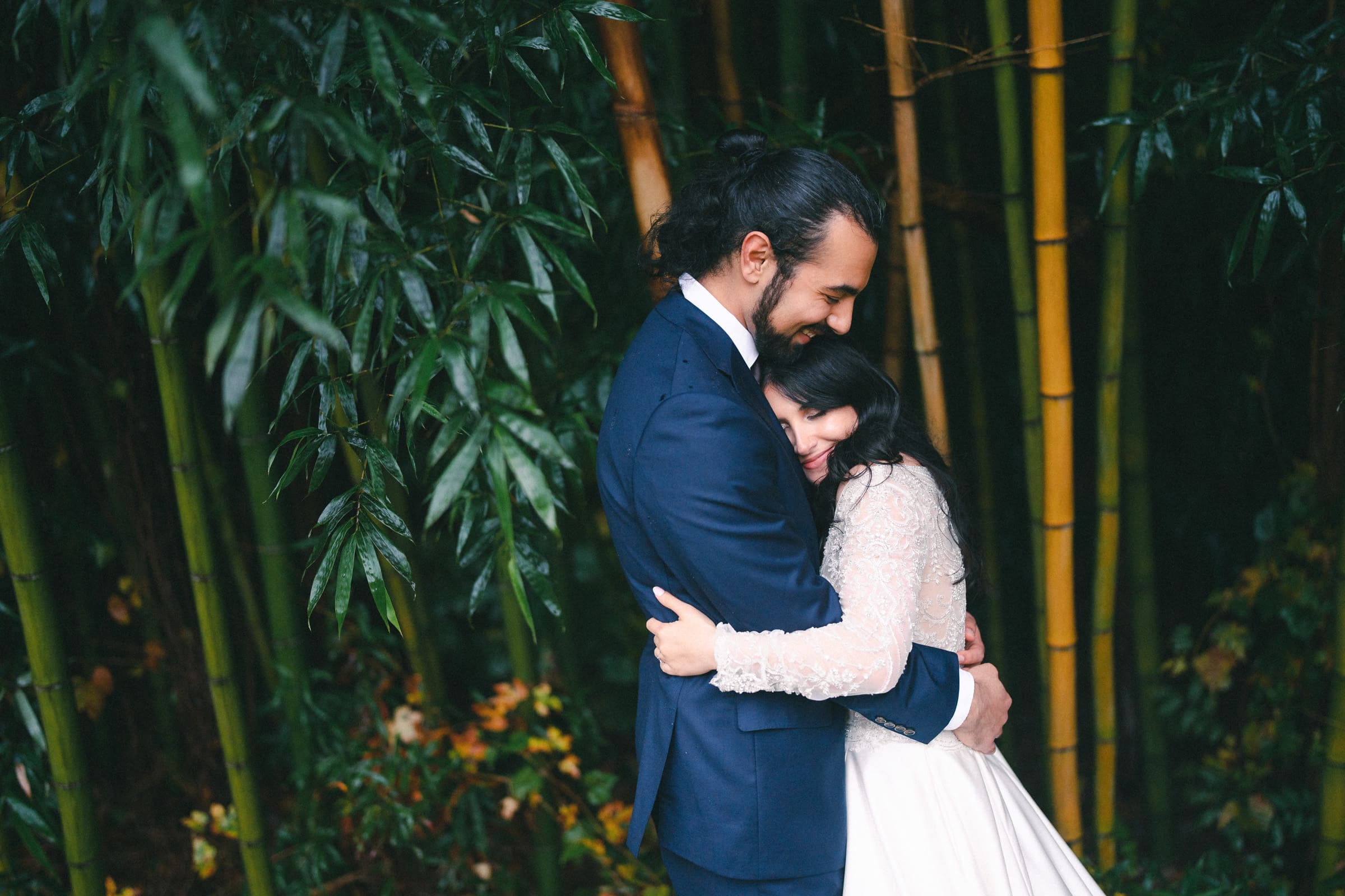 A couple embraces tenderly amidst the serene beauty of a lush forest, reminiscent of a Graylyn Estate wedding. The man dons a navy suit, while the woman wears a white dress with lace sleeves. Tall green bamboo stalks surround them, enhancing the intimate atmosphere.