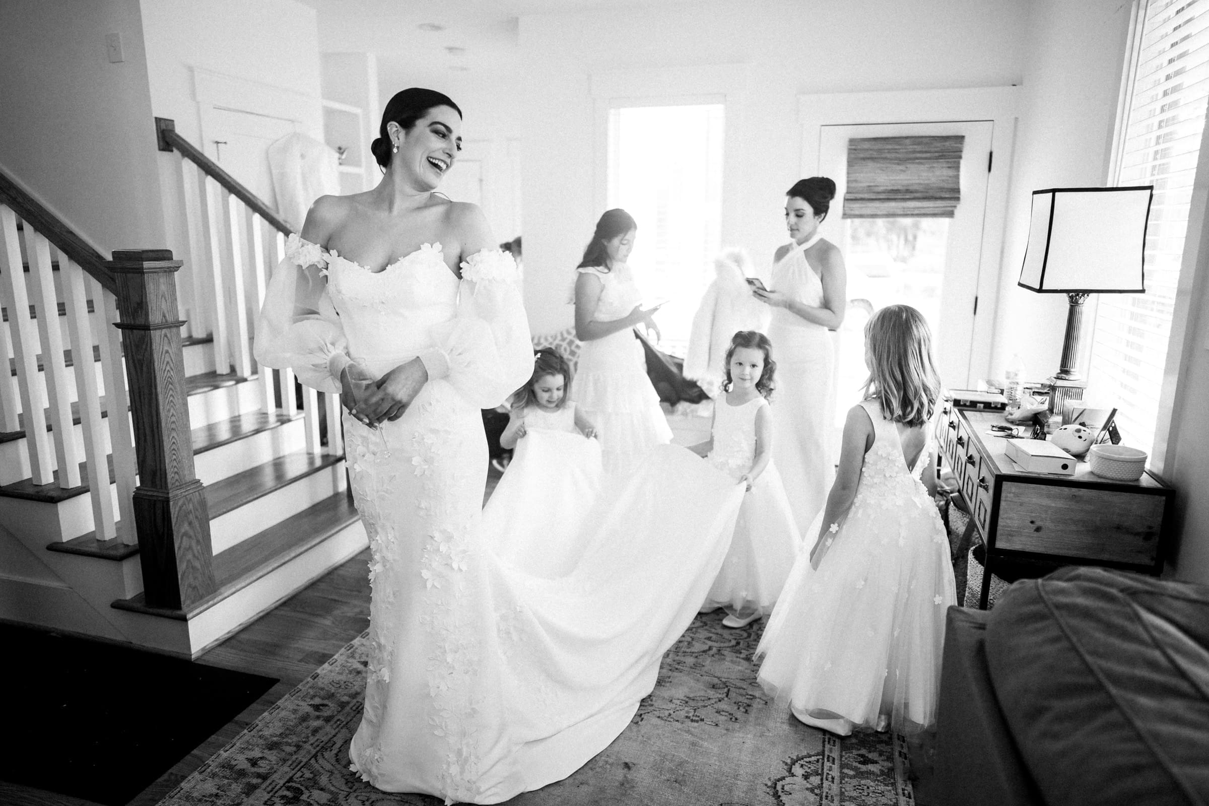 In a bright room at the Melrose Knitting Mill wedding, a bride in an off-shoulder gown smiles as bridesmaids and flower girls adjust her train. The scene is lively and elegant, with large windows and a grand staircase enhancing the ambiance.