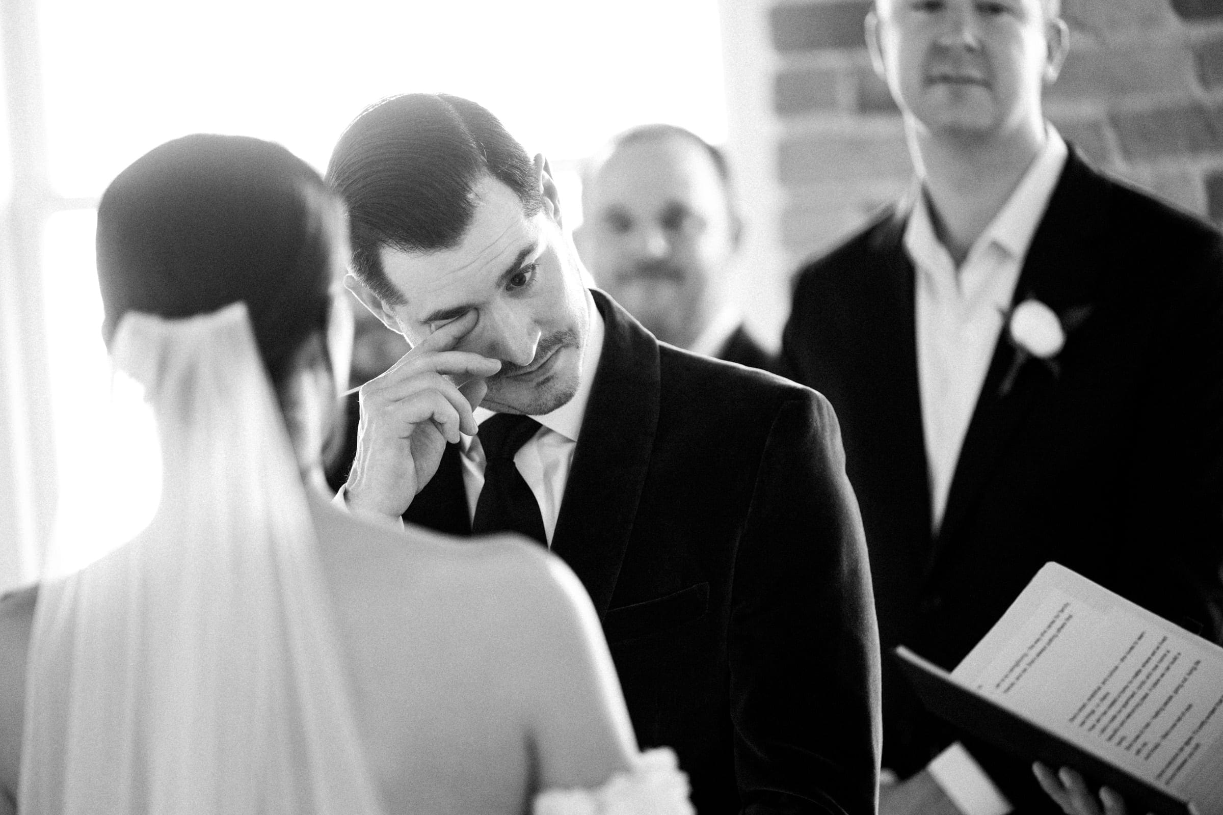 At a Melrose Knitting Mill wedding, a groom in a suit wipes a tear from his eye during the ceremony. The bride, in a white dress with a veil, faces him as two groomsmen stand nearby. The scene is filled with emotion and intimacy.