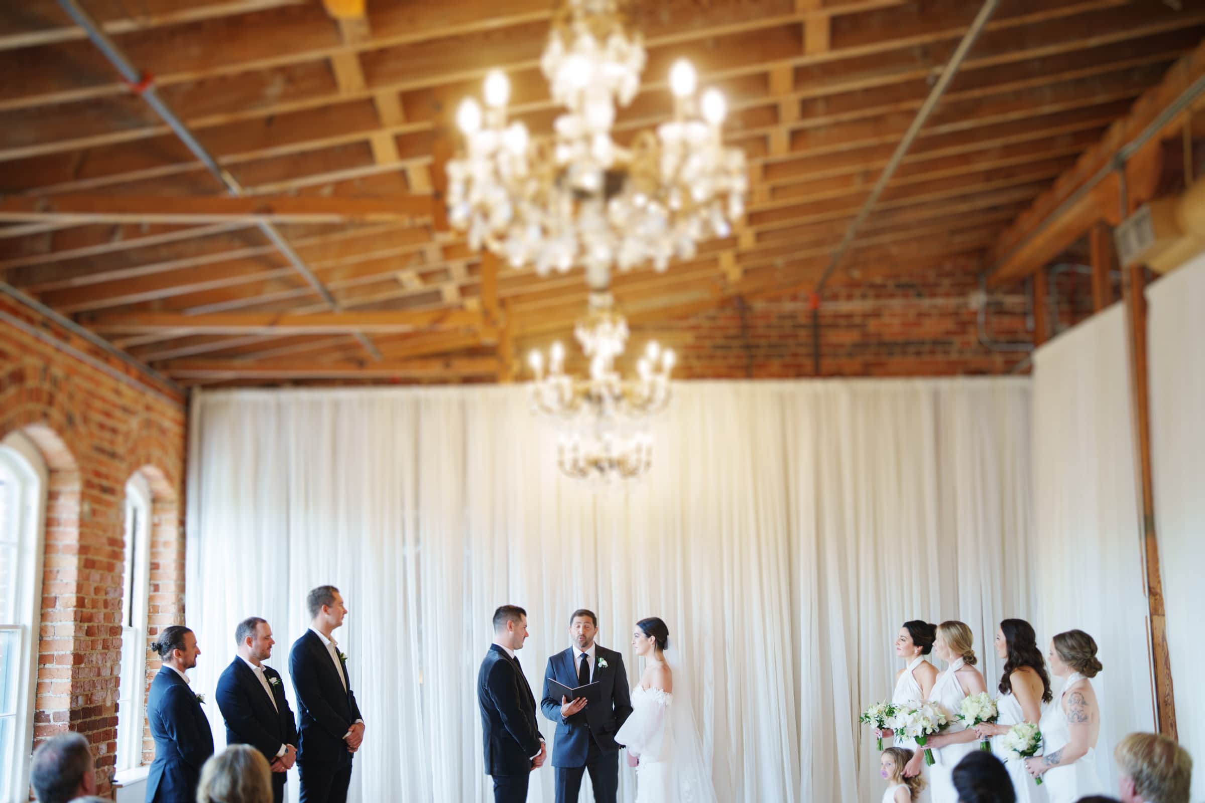 A wedding ceremony unfolds at the Melrose Knitting Mill, a rustic venue with exposed brick walls and chandeliers. The bride and groom stand at the altar with the officiant, while the bridal party, in pastel dresses and suits, stands elegantly on either side.