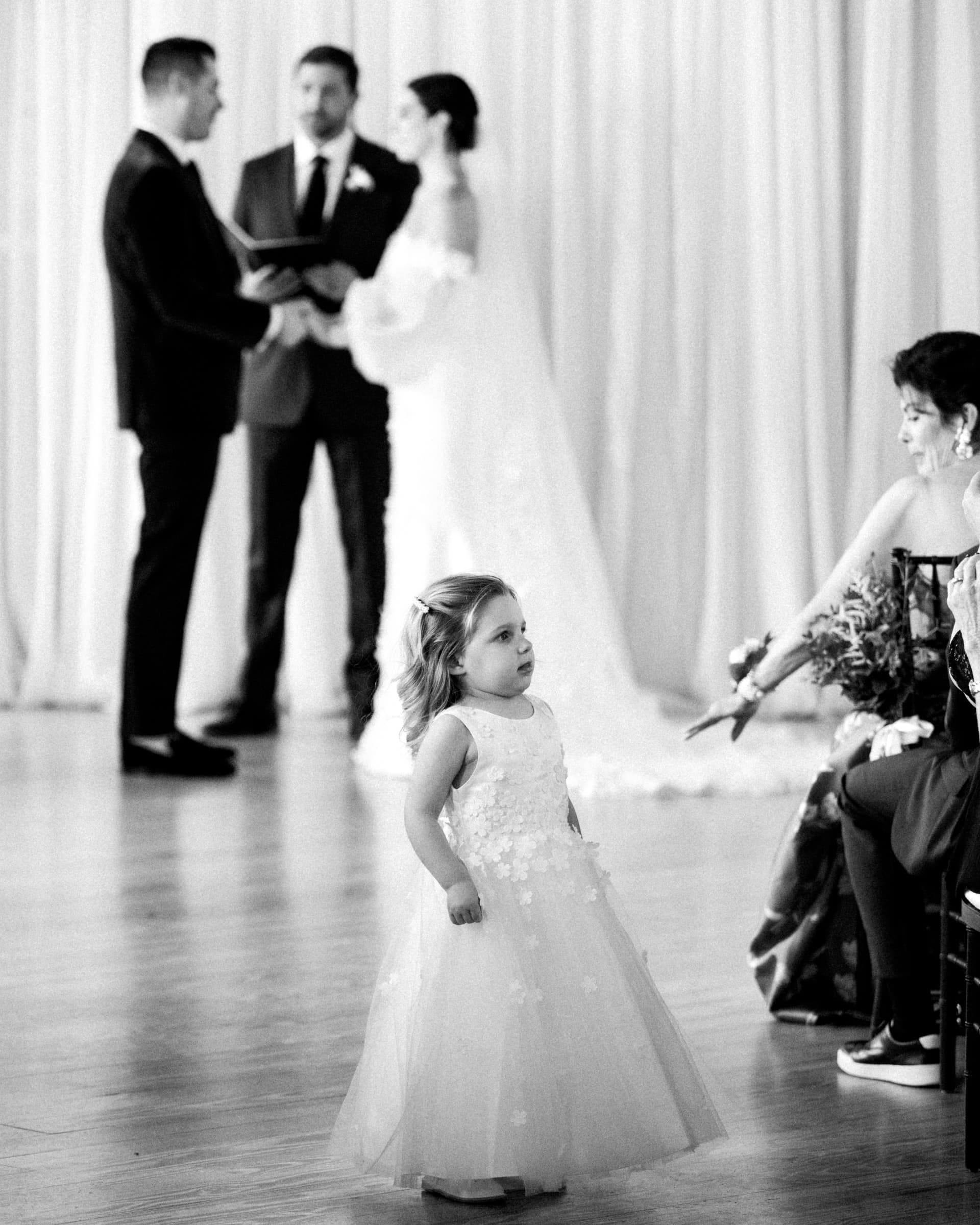 A young girl in a white dress stands at a Melrose Knitting Mill wedding. In the background, a couple exchanges vows with an officiant, while guests sit to the side. The image is in black and white, capturing timeless elegance and charm.
