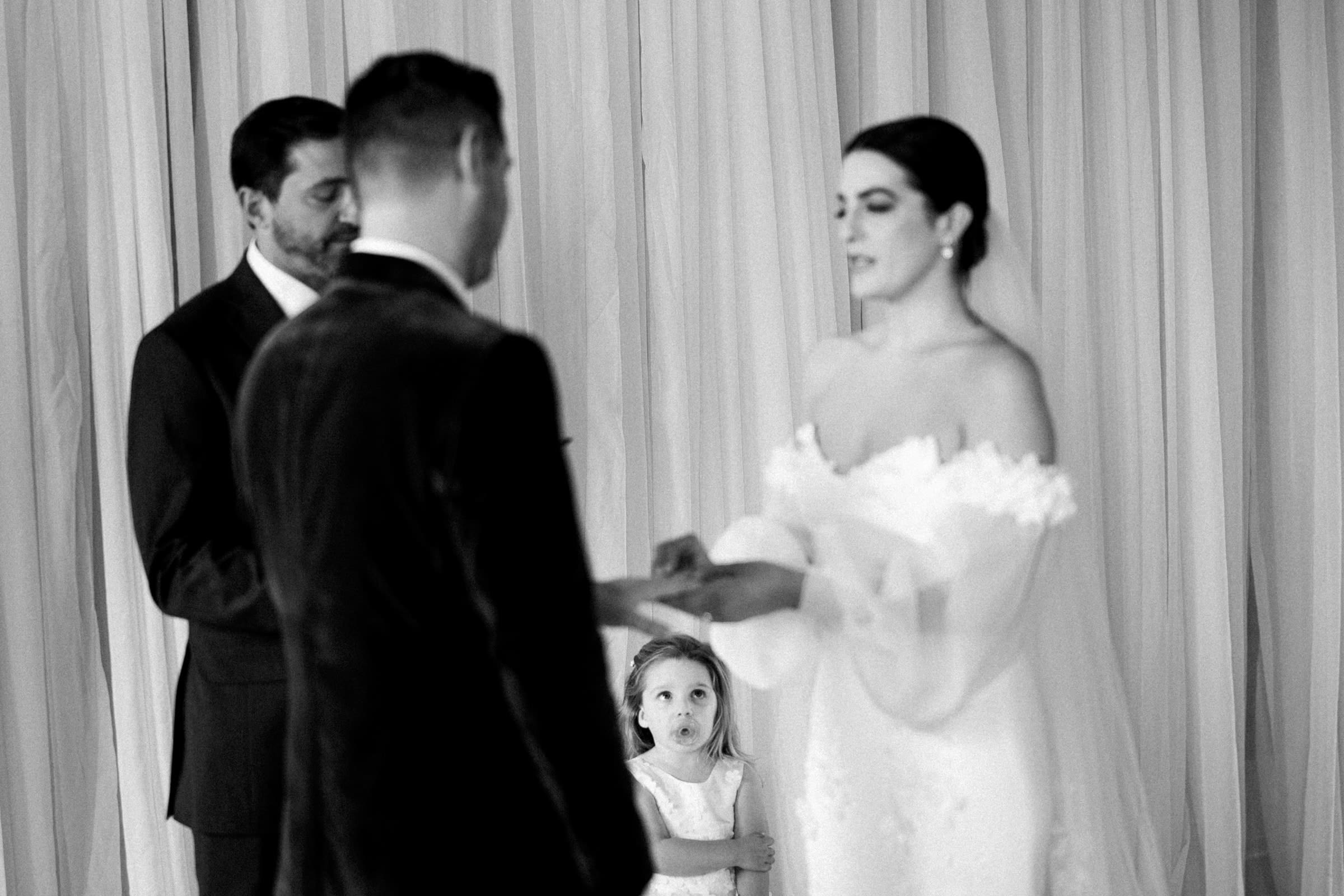 A black and white image captures a Melrose Knitting Mill wedding. Two people stand facing each other, exchanging rings as a person officiates beside them. A young child looks up at the couple. The elegant setting, with draped curtains, adds to the timeless charm of this special moment.