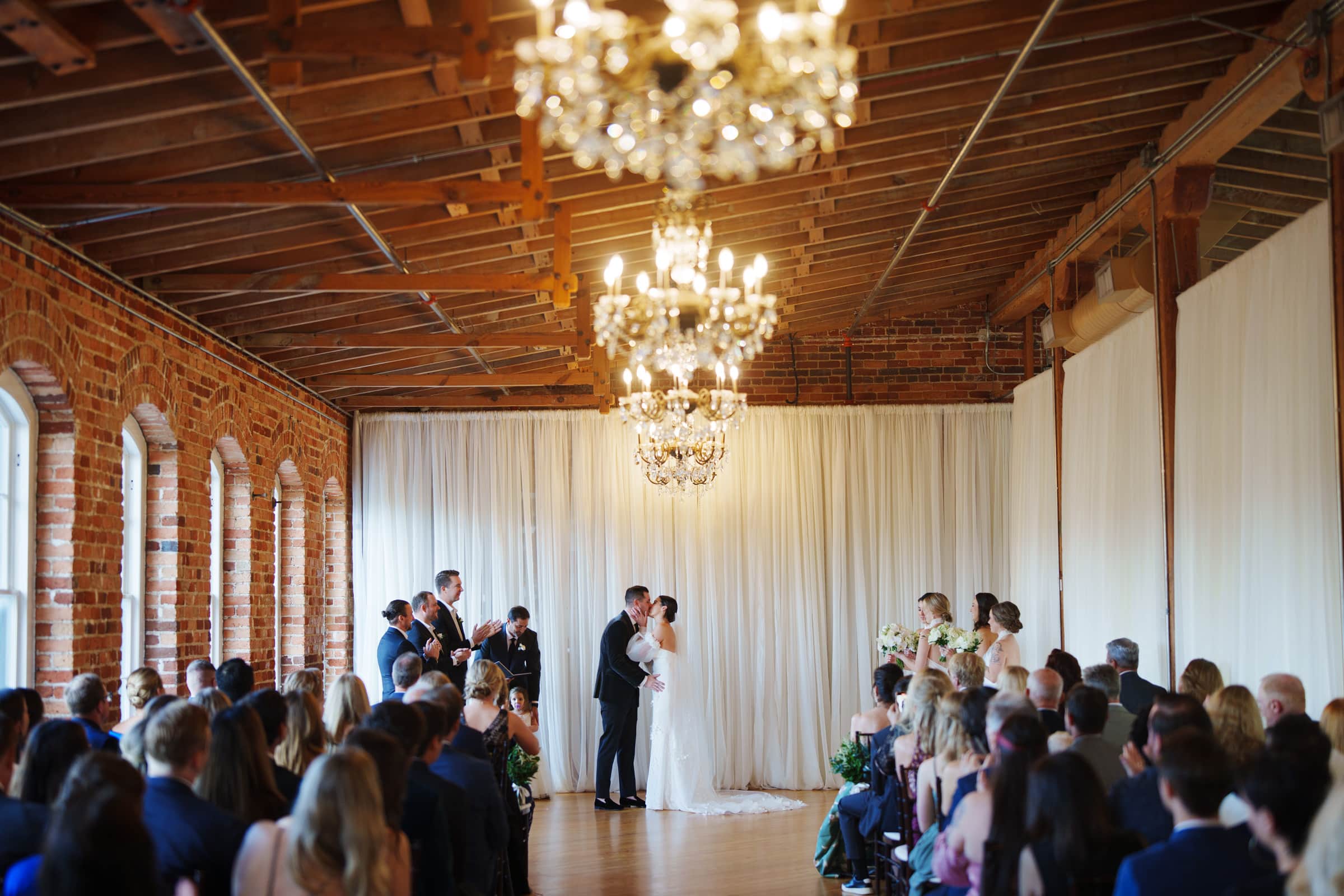 A Melrose Knitting Mill wedding unfolds beautifully in the rustic venue adorned with exposed brick and chandeliers. At the altar, the couple shares a kiss amidst their elegantly dressed bridal party, as guests are seated in awe, witnessing this intimate moment.