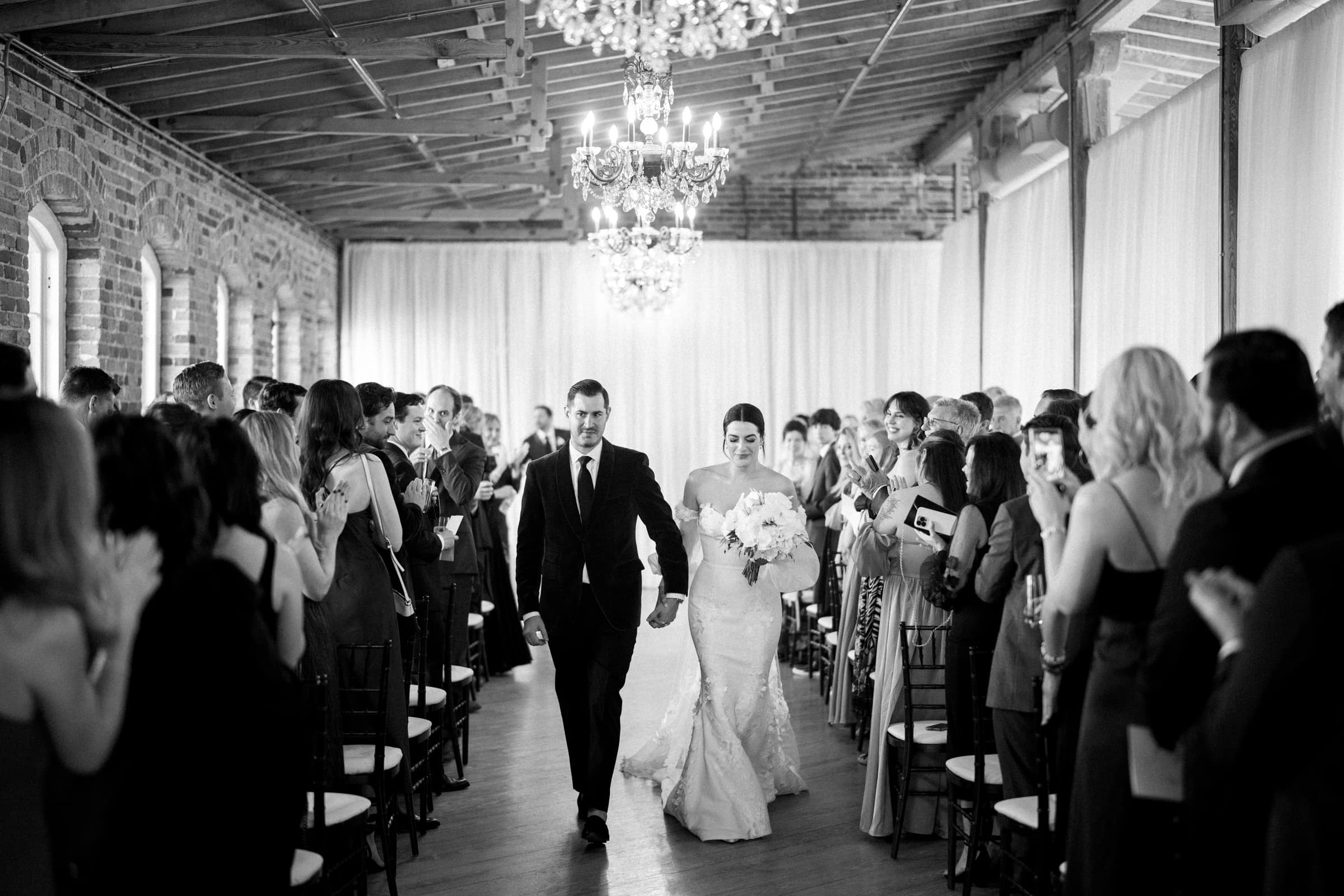 A bride and groom walk down the aisle at their Melrose Knitting Mill wedding, smiling and hand in hand. Guests on both sides are seated and clapping. The elegant room, with chandeliers and exposed brick walls, is beautifully captured in a timeless black-and-white scene.