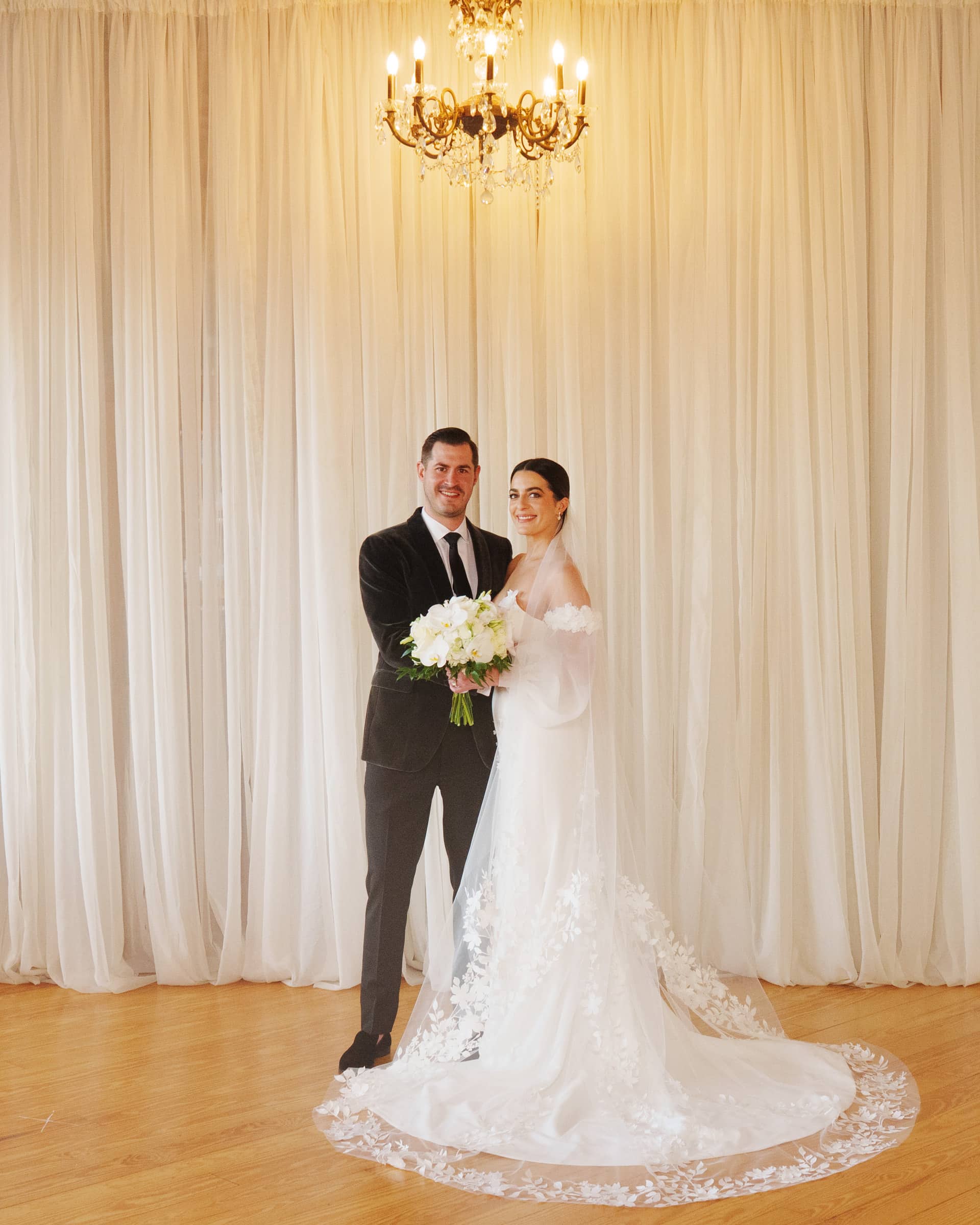 A bride and groom stand together at a Melrose Knitting Mill wedding, the warmly lit room illuminated by a chandelier above. The bride wears a long white gown with lace details, holding a bouquet of white flowers, while the groom is in a dark suit. A sheer white curtain serves as the backdrop.