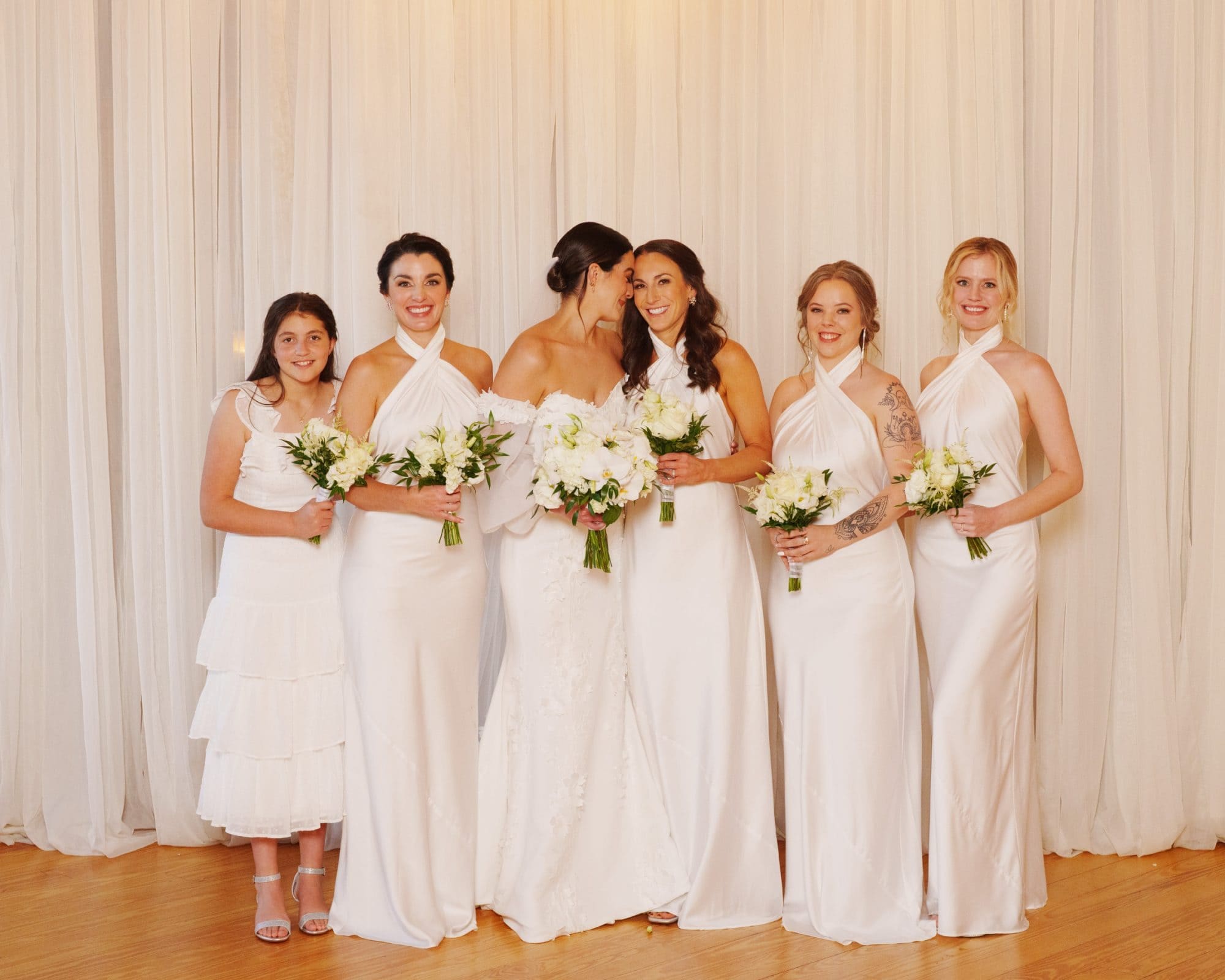 At a picturesque Melrose Knitting Mill wedding, the bride stands with her five bridesmaids, all dressed in elegant white and holding stunning bouquets. Positioned before a light beige curtain backdrop, they smile radiantly at the camera.