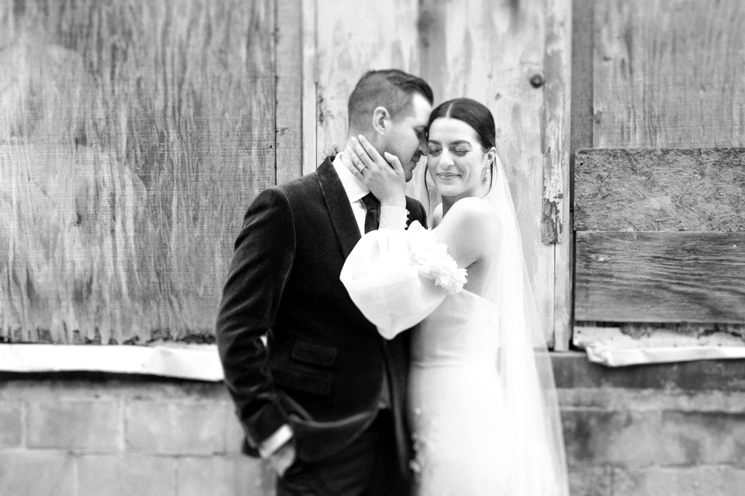 A black and white photo captures a bride and groom at their Melrose Knitting Mill wedding, standing closely against a rustic wooden background. The bride, in her white dress and veil, gently holds the groom's face as he smiles with closed eyes in his dark suit.