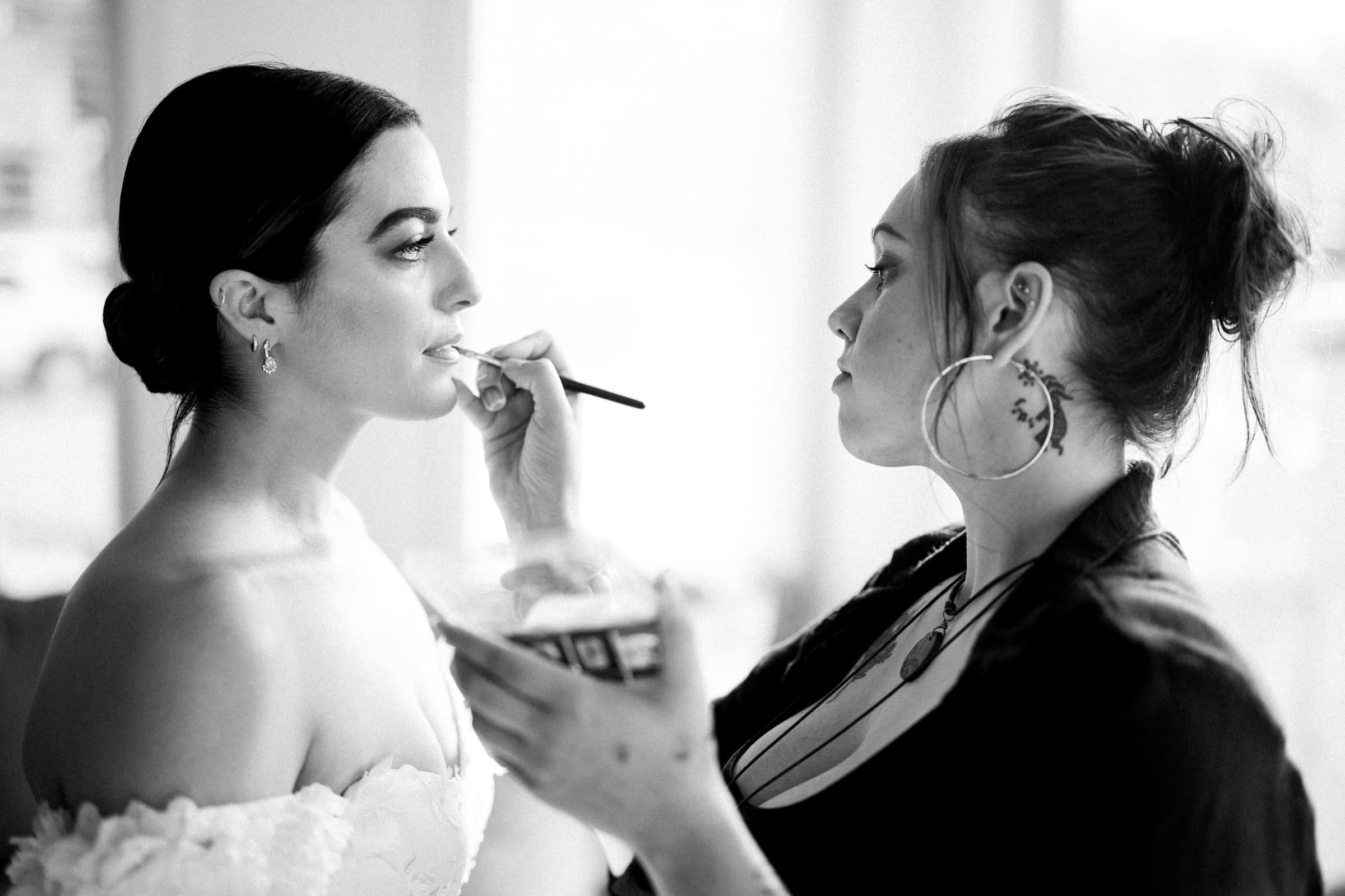 At a Melrose Knitting Mill wedding, a makeup artist applies lipstick to a woman in a bridal gown. Her hair is styled in an elegant updo as the artist, focused and poised, holds a brush and palette in the softly lit room. The black and white image captures timeless elegance amidst their preparations.