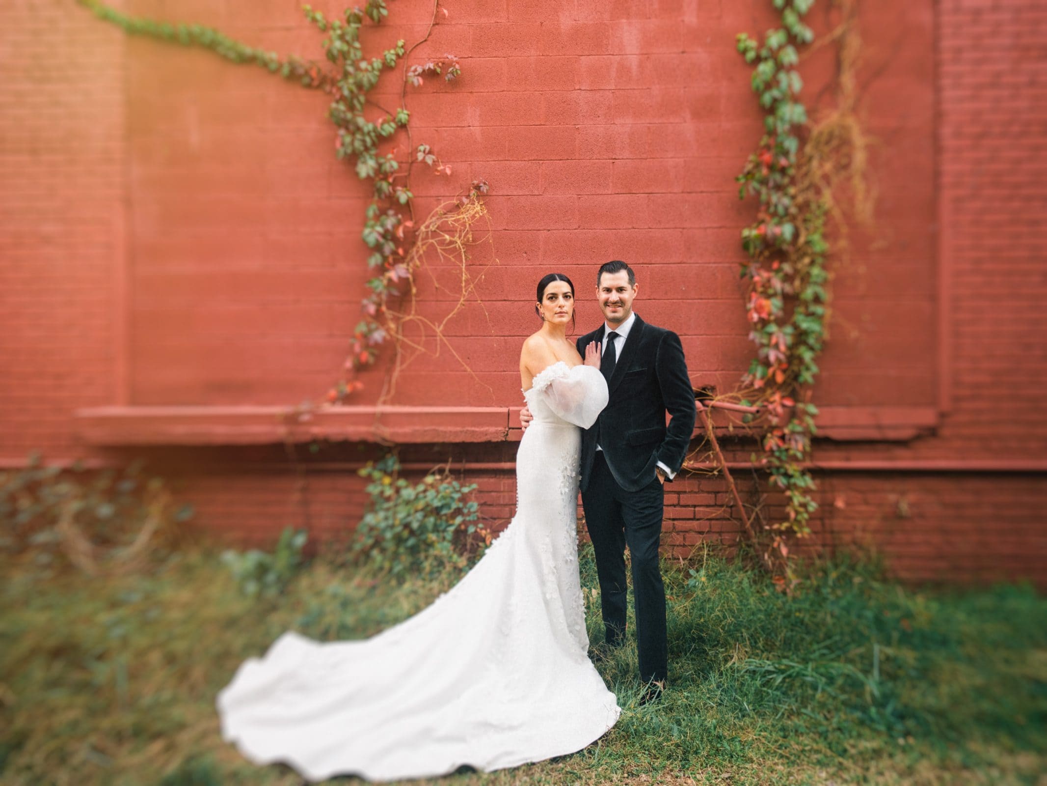 The bride in a long white gown and groom in a black tuxedo stand together at their Melrose Knitting Mill wedding, framed by a red brick wall adorned with green and red ivy. Holding her bouquet, they both gaze towards the camera, set against a soft, blurry backdrop.