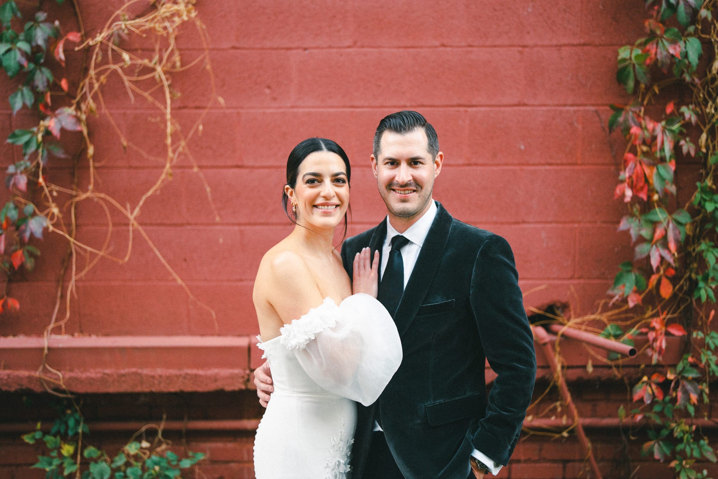 A bride and groom stand close together, smiling, in formal attire at a Melrose Knitting Mill wedding. The bride wears an off-shoulder white dress, and the groom is in a black suit. They are in front of a red brick wall adorned with green and red ivy.