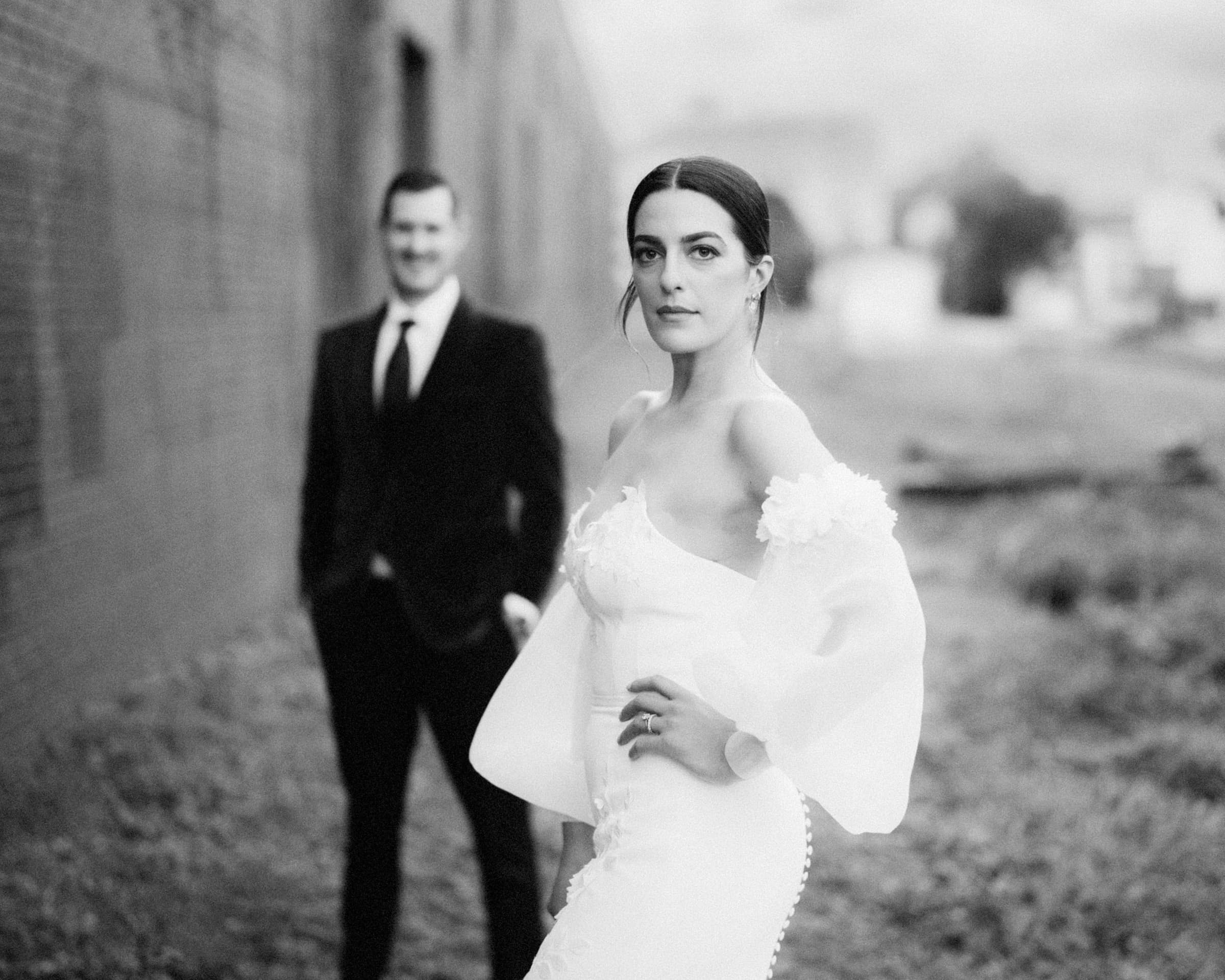 A black-and-white photo captures a bride in a wedding dress, gazing at the camera amidst the industrial charm of Melrose Knitting Mill. In focus, she stands elegantly as a man in a suit is blurred in the background, adding depth to this timeless moment.