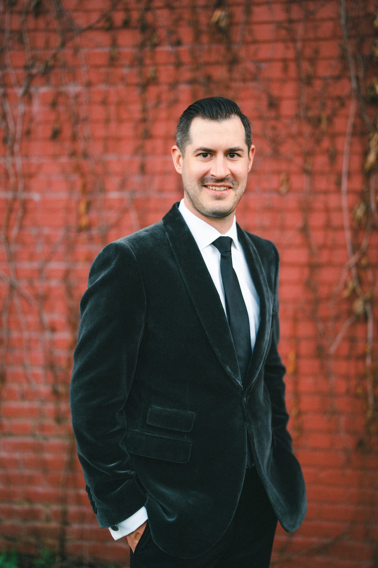 A man in a black suit and tie stands smiling in front of a red brick wall with leafy vines, capturing the timeless elegance of a Melrose Knitting Mill wedding.