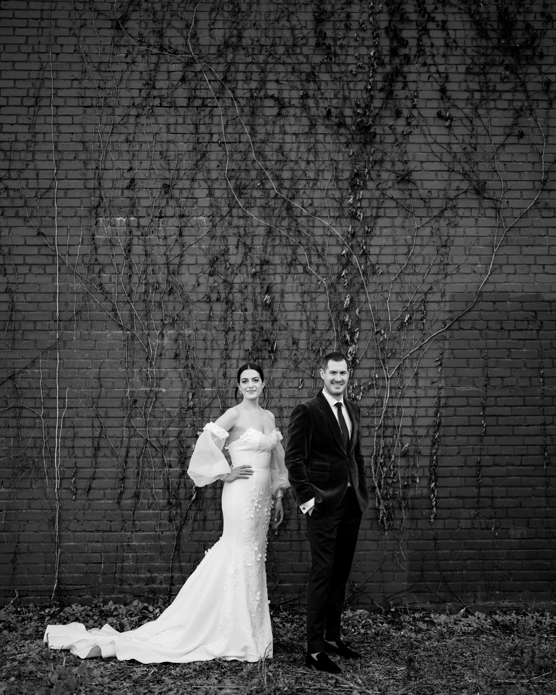 In a timeless black-and-white photo, a bride and groom stand before a vine-draped brick wall, reminiscent of a Melrose Knitting Mill wedding. The bride graces an elegant white gown, while the groom dons a dark suit. United in hand, they share subtle smiles.