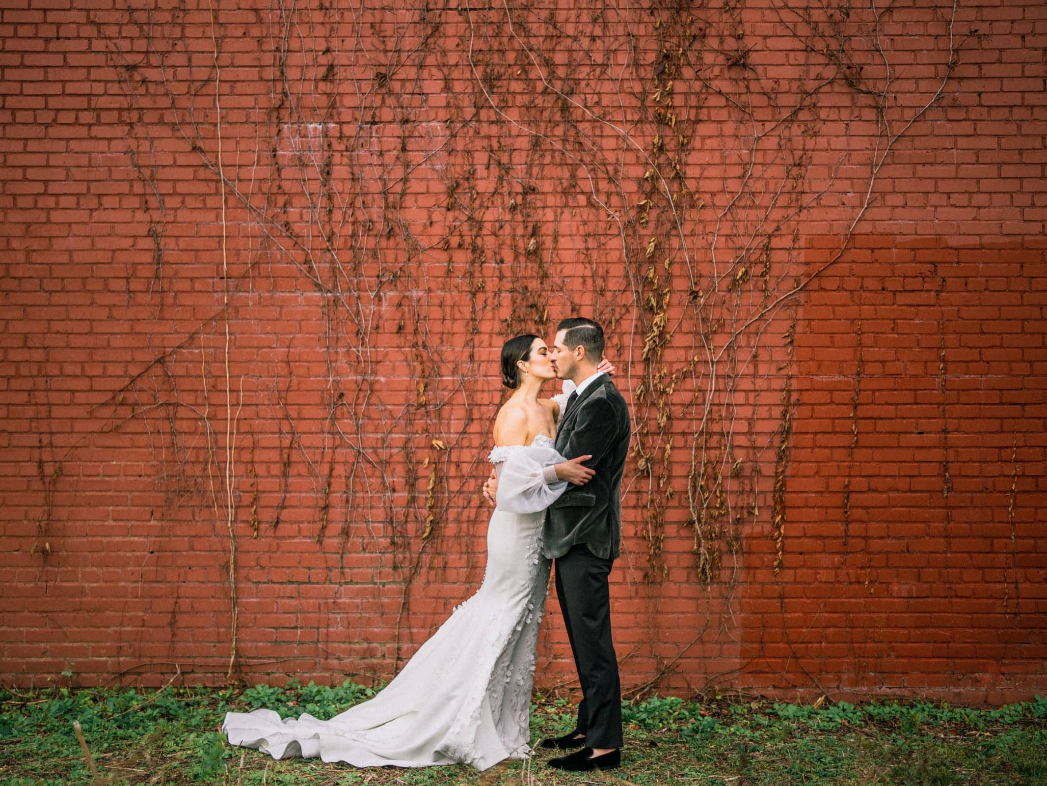 A couple shares a kiss in front of a brick wall with climbing vines, embodying the charm of a Melrose Knitting Mill wedding. The bride stuns in her long, white gown while the groom complements her beautifully in his dark suit. Green grass lines the bottom of this picturesque backdrop.