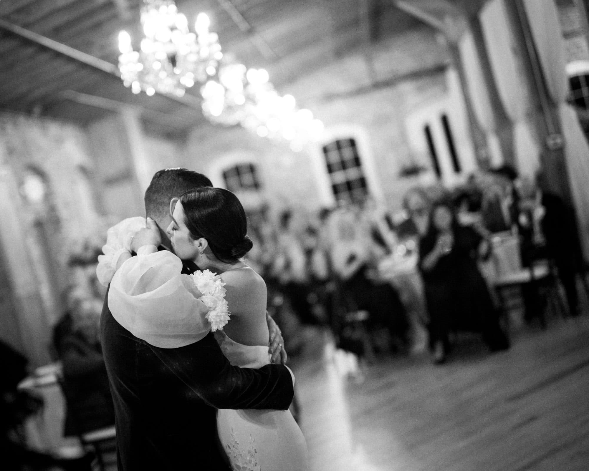 A couple embraces on the dance floor during a Melrose Knitting Mill wedding. The bride in her white gown and the groom in his dark suit sway under the glow of chandeliers. Guests enjoy the softly lit, rustic venue, adding to the enchanting ambiance of this memorable occasion.
