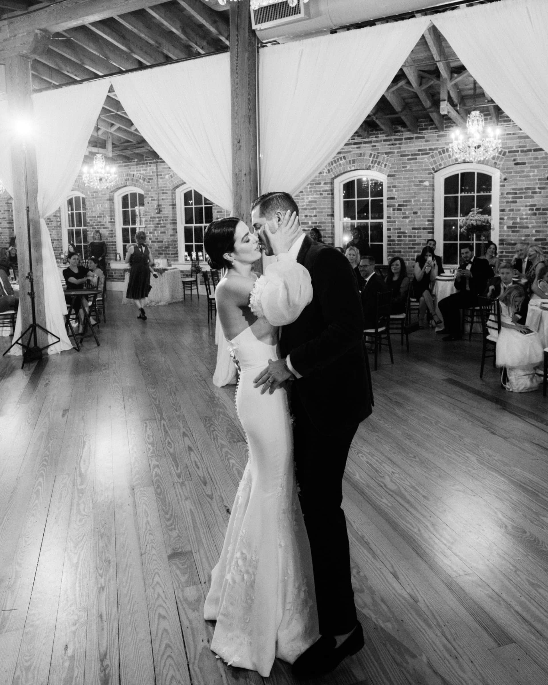 A bride and groom share a romantic dance in the spacious, elegant Melrose Knitting Mill venue with wooden floors and brick walls. Draped curtains hang from the ceiling, and guests sit at tables in the background, watching the couple.