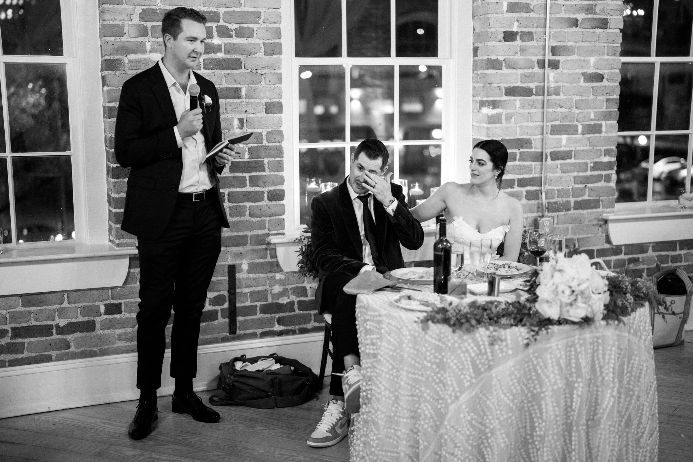 In a black-and-white photo from a Melrose Knitting Mill wedding, a man in a suit gives a heartfelt speech into the microphone. The seated groom wipes his eye beside the bride at their flower-adorned table. They are indoors, surrounded by brick walls and large windows.