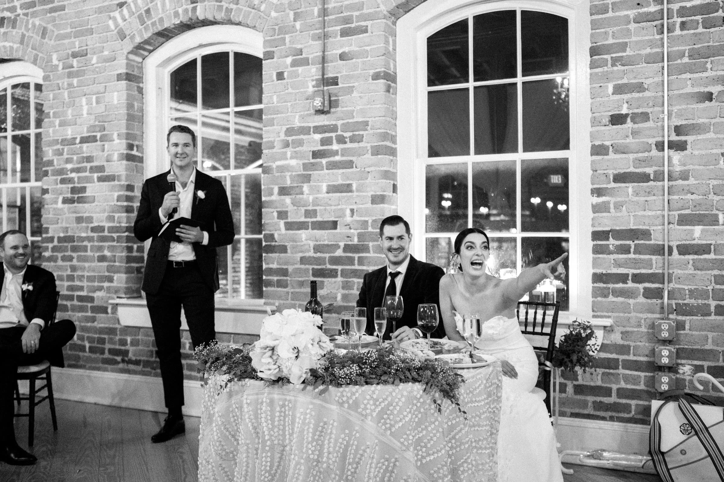 At a charming Melrose Knitting Mill wedding, the bride and groom sit at a decorated table in front of a brick wall with arched windows. A man stands giving a speech, while the bride gestures enthusiastically. Two other guests are seated nearby, all in a warmly lit room.