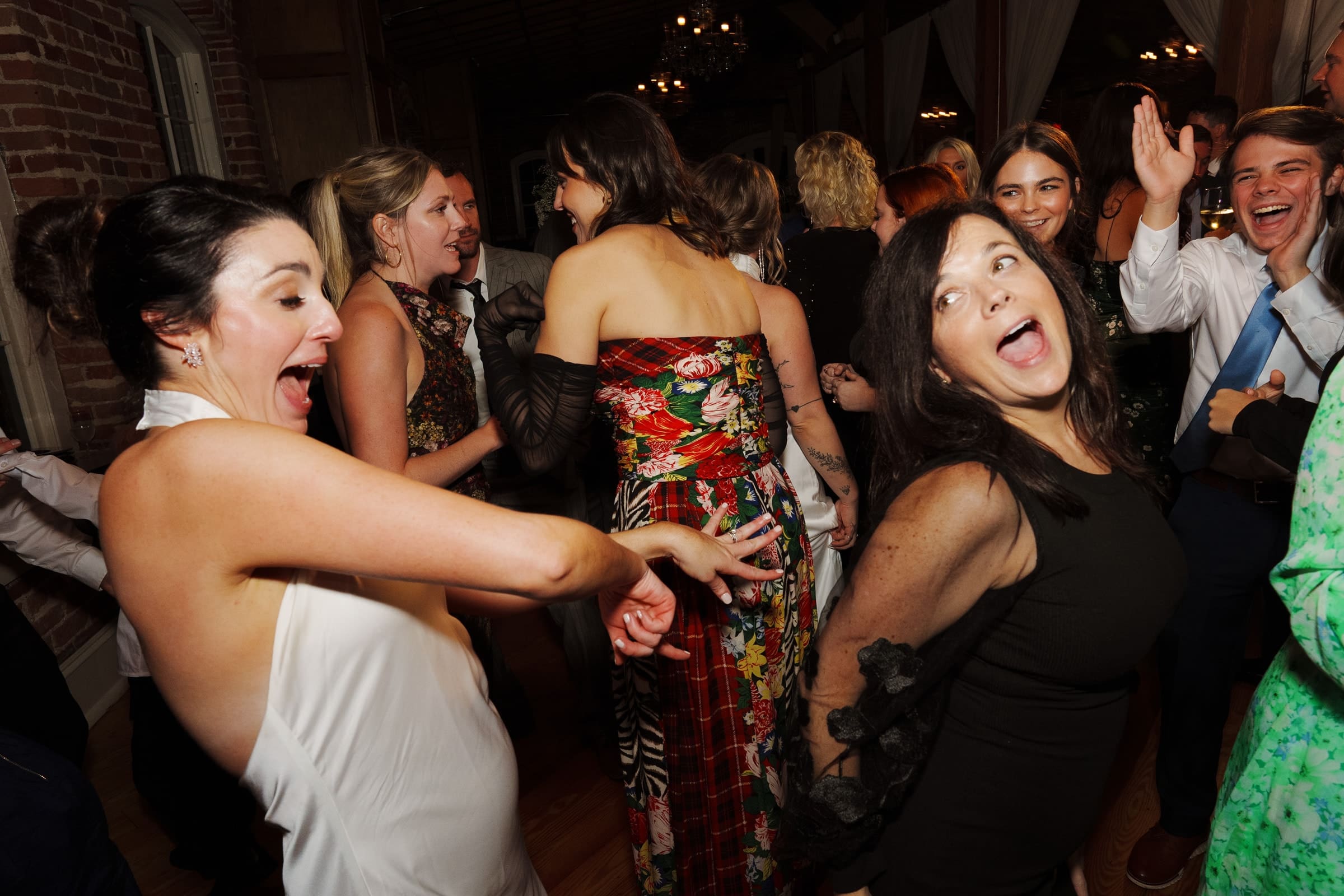 A group of people dancing energetically at a lively indoor event at the Melrose Knitting Mill wedding. They are dressed in formal and colorful attire, with expressions of joy and excitement on their faces. The warmly lit setting features brick walls and chandeliers, enhancing the festive atmosphere.