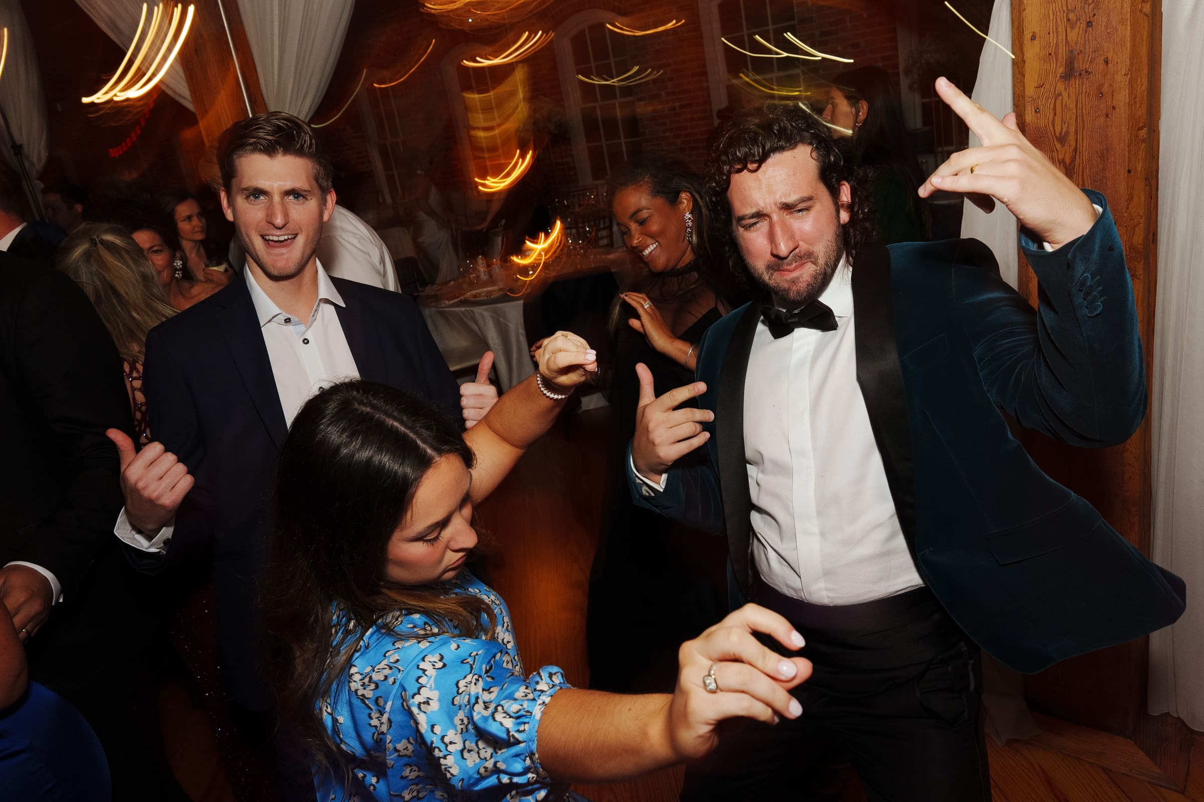 A lively group of people dancing at a party in Melrose Knitting Mill, with wooden floors and draped walls. A man in a blue suit and another in a tuxedo strike expressive dance poses, while a woman in a blue patterned dress dances energetically beside them.