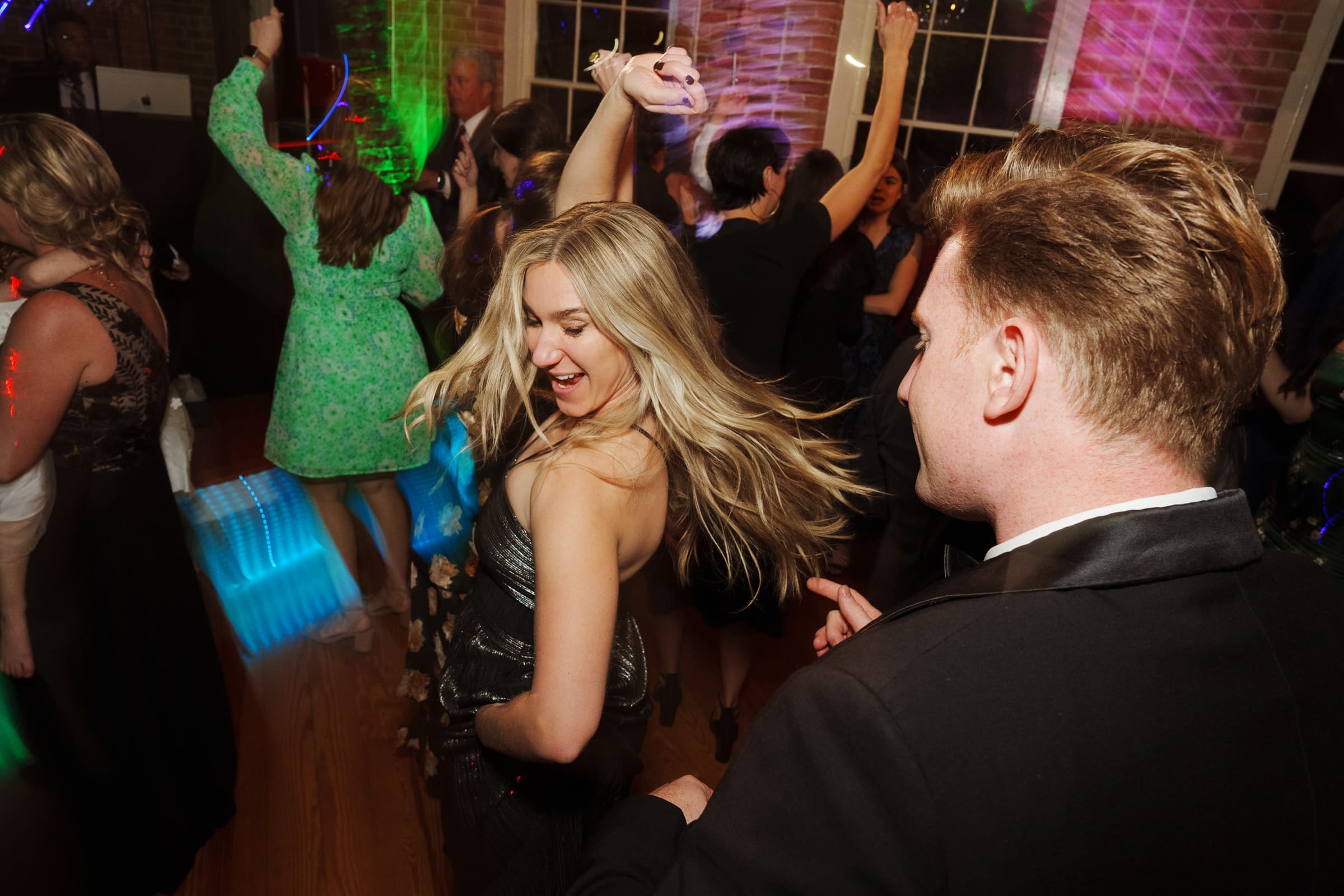 At a vibrant Melrose Knitting Mill wedding, people dance energetically. A woman in a sparkling dress is at the center, smiling with her hair flowing. Colorful lights and a lively atmosphere fill the room, with others dancing and reveling in the joyous celebration.