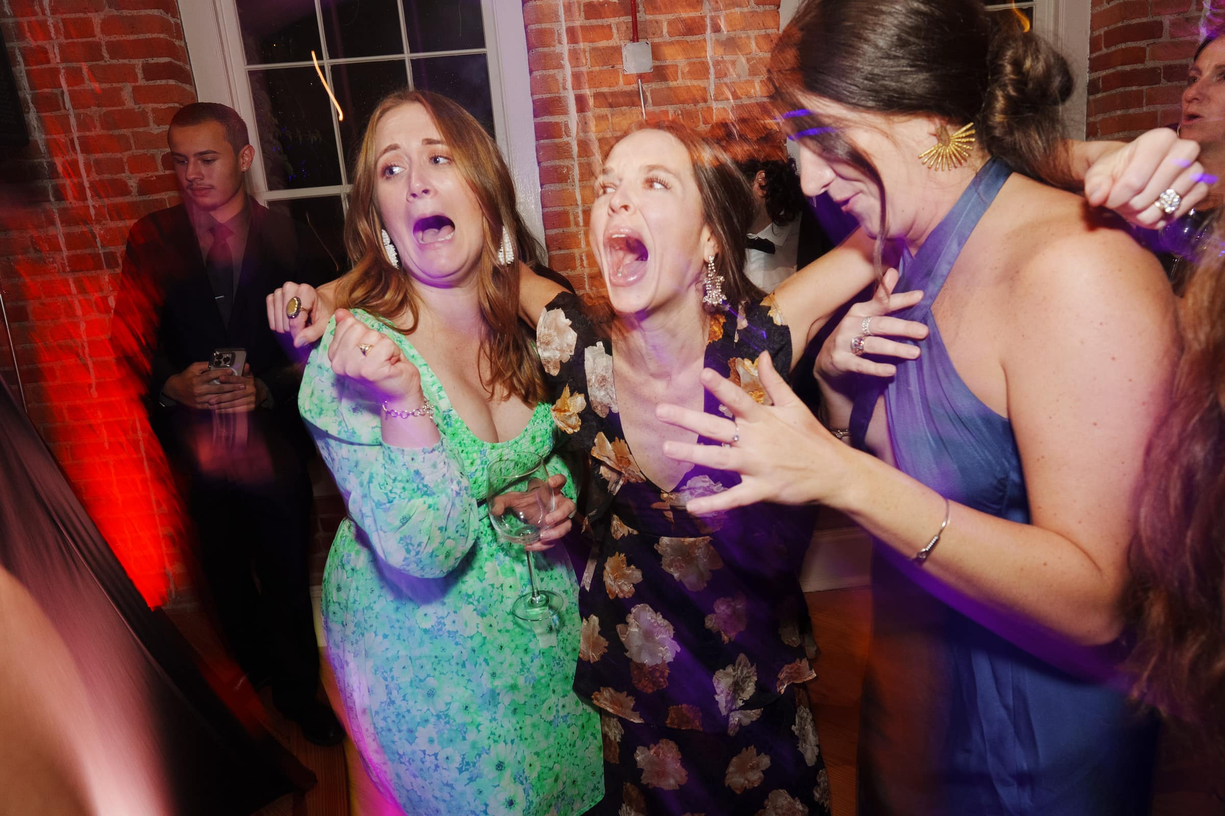 Three women in colorful dresses are energetically singing with expressions of enthusiasm and joy at a lively Melrose Knitting Mill wedding inside a room with brick walls. A man in the background is observing while holding a drink.