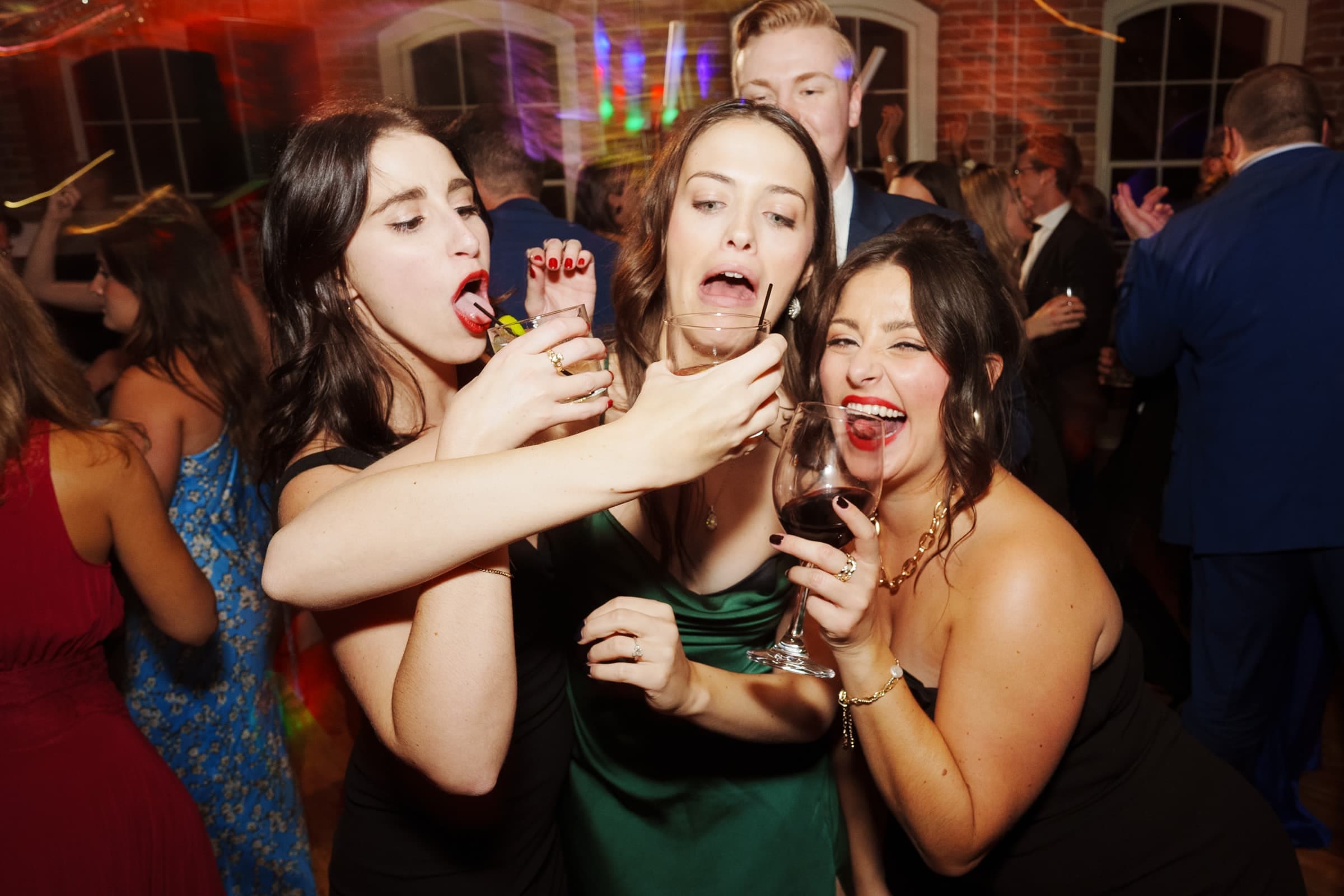 Three women in party dresses hold wine glasses and make playful expressions, with one pretending to scream and others laughing at the vibrant Melrose Knitting Mill wedding. People are dancing in the background of a lively, colorful room.