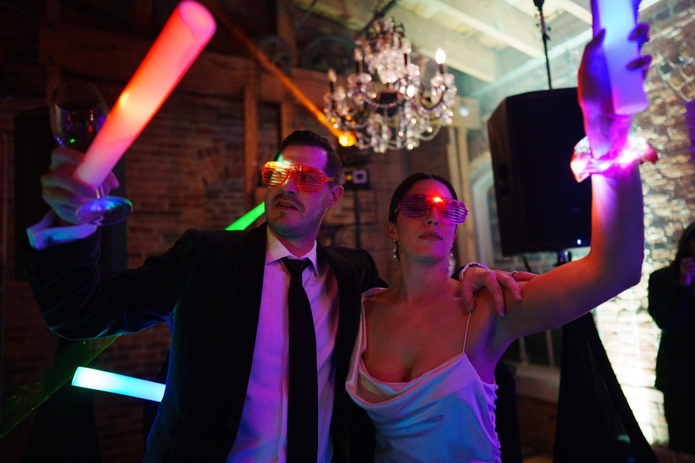 A man in a suit and a woman in a white dress pose at the Melrose Knitting Mill wedding, both wearing glowing shutter shades and holding colorful light sticks. A chandelier and exposed brick wall enhance the festive atmosphere.