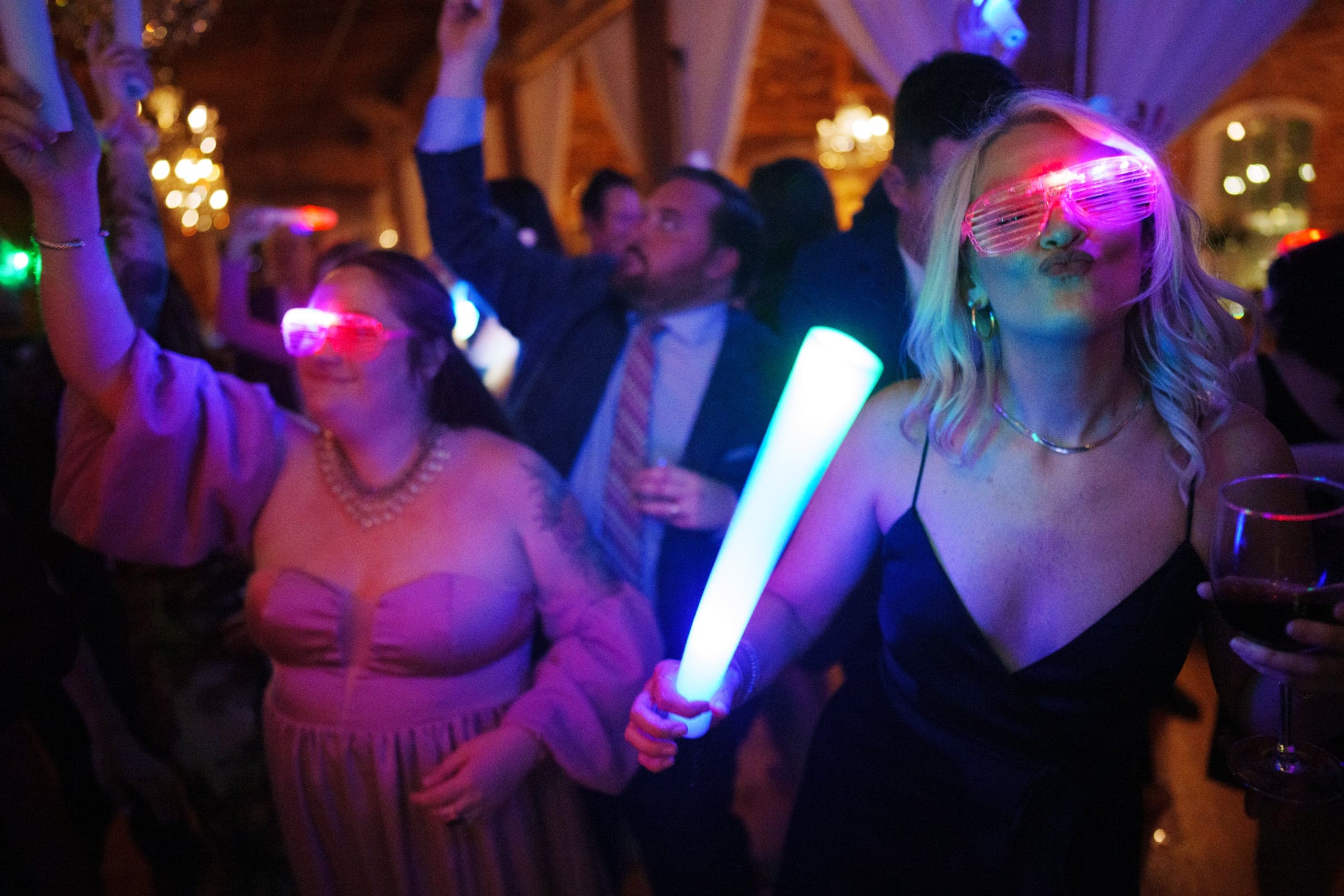 A lively party scene at the Melrose Knitting Mill wedding, with people dancing and smiling. Two individuals in the foreground wear glowing glasses and hold illuminated sticks. The warmly lit room, adorned with chandeliers, enhances the joyous atmosphere.