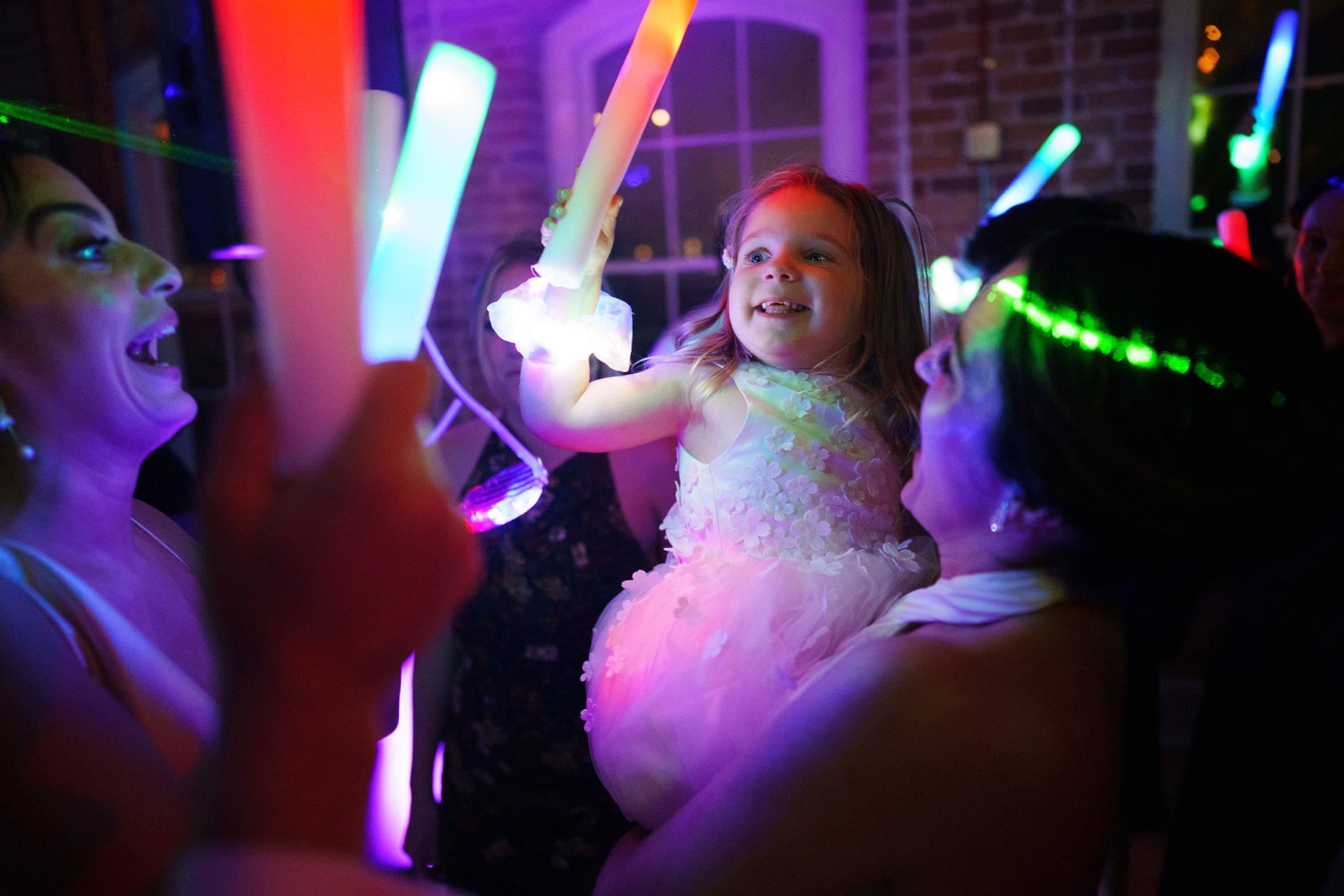 A joyful moment unfolds at a Melrose Knitting Mill wedding, where a young girl in a white dress is held up, smiling and holding a glowing light stick. Colorful lights illuminate the scene, with guests around her reveling in the celebration.