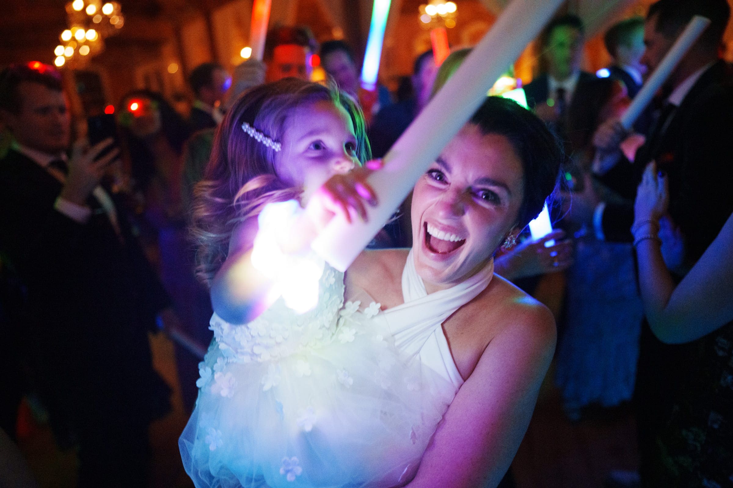 At a lively Melrose Knitting Mill wedding, a woman in a white dress joyfully holds a young girl in a flower dress. Both are smiling and waving brightly lit glow sticks as they join the celebration, surrounded by colorful lights and cheerful guests.