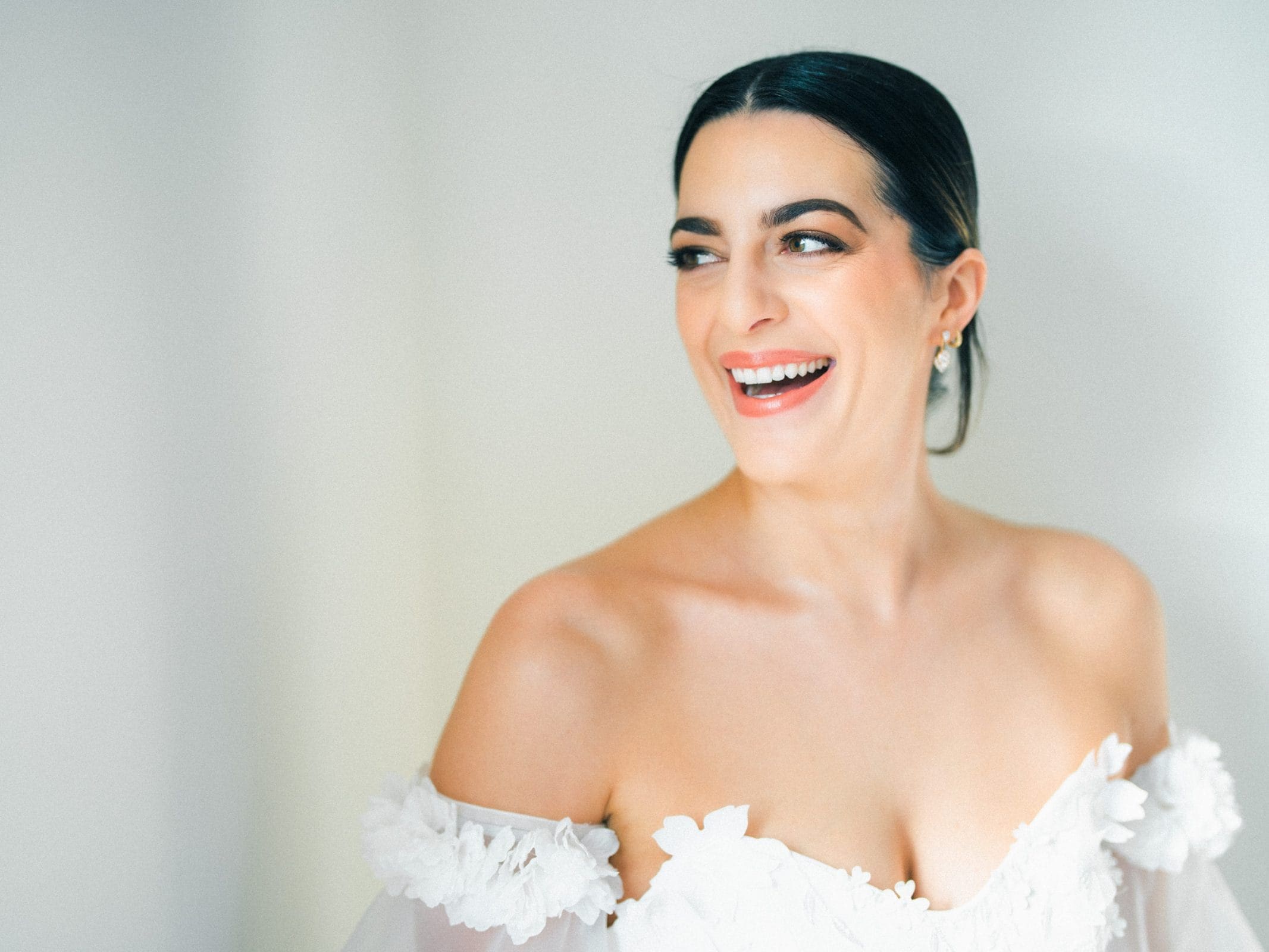 A woman with dark hair pulled back smiles broadly, wearing an off-the-shoulder white dress adorned with floral details at a Melrose Knitting Mill wedding. The background is softly lit, creating a warm and elegant atmosphere.