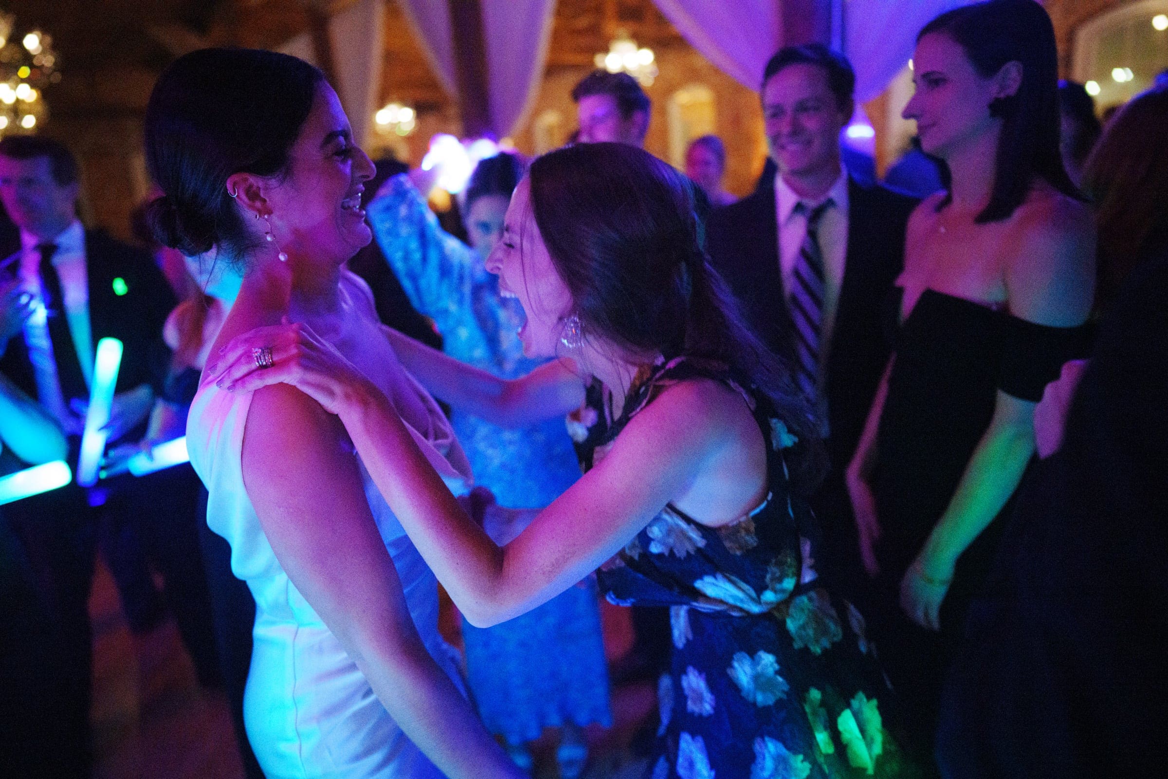 Two women in party attire share a joyful moment on the dance floor of the Melrose Knitting Mill wedding, surrounded by friends dressed formally. The scene is vibrant, with colorful lights creating a lively atmosphere.