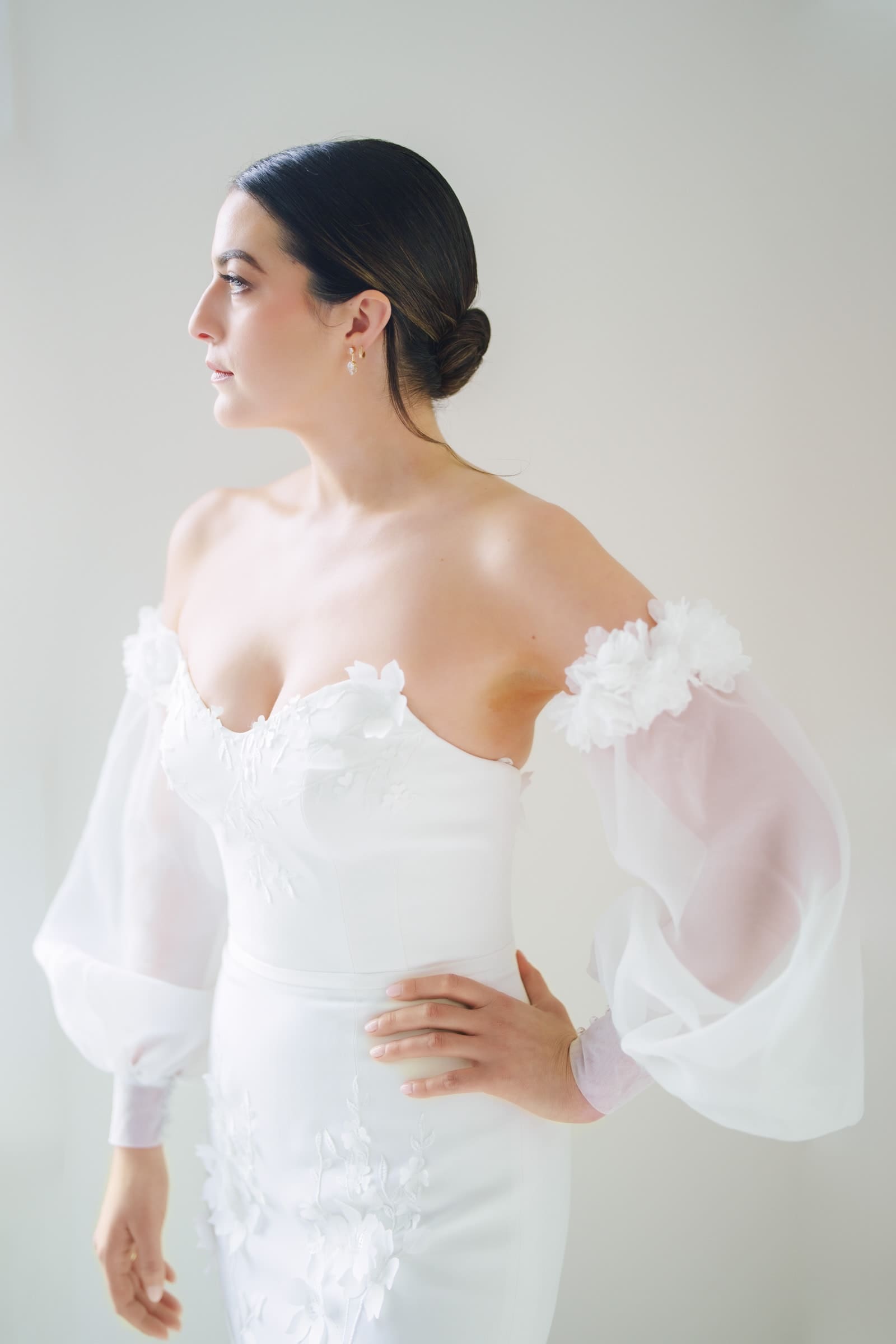 A woman in an elegant off-the-shoulder white wedding dress, adorned with floral designs and sheer long sleeves, stands in profile. Her dark hair is styled in a low bun as she gazes thoughtfully into the distance, embodying the timeless grace of a Melrose Knitting Mill wedding. 
