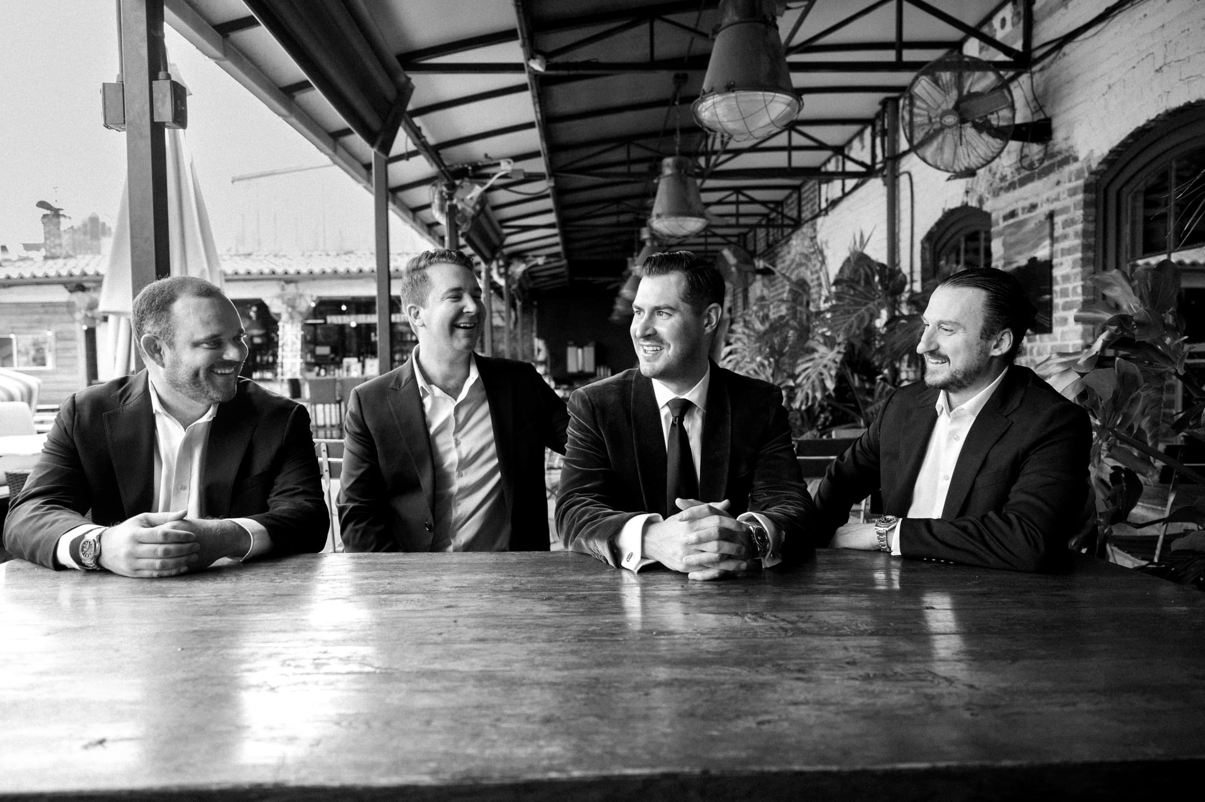 Four men in suits are sitting at a wooden table, laughing and engaged in conversation at a Melrose Knitting Mill wedding. They are in a covered outdoor area with large windows and industrial-style lighting, setting a relaxed and cheerful mood.