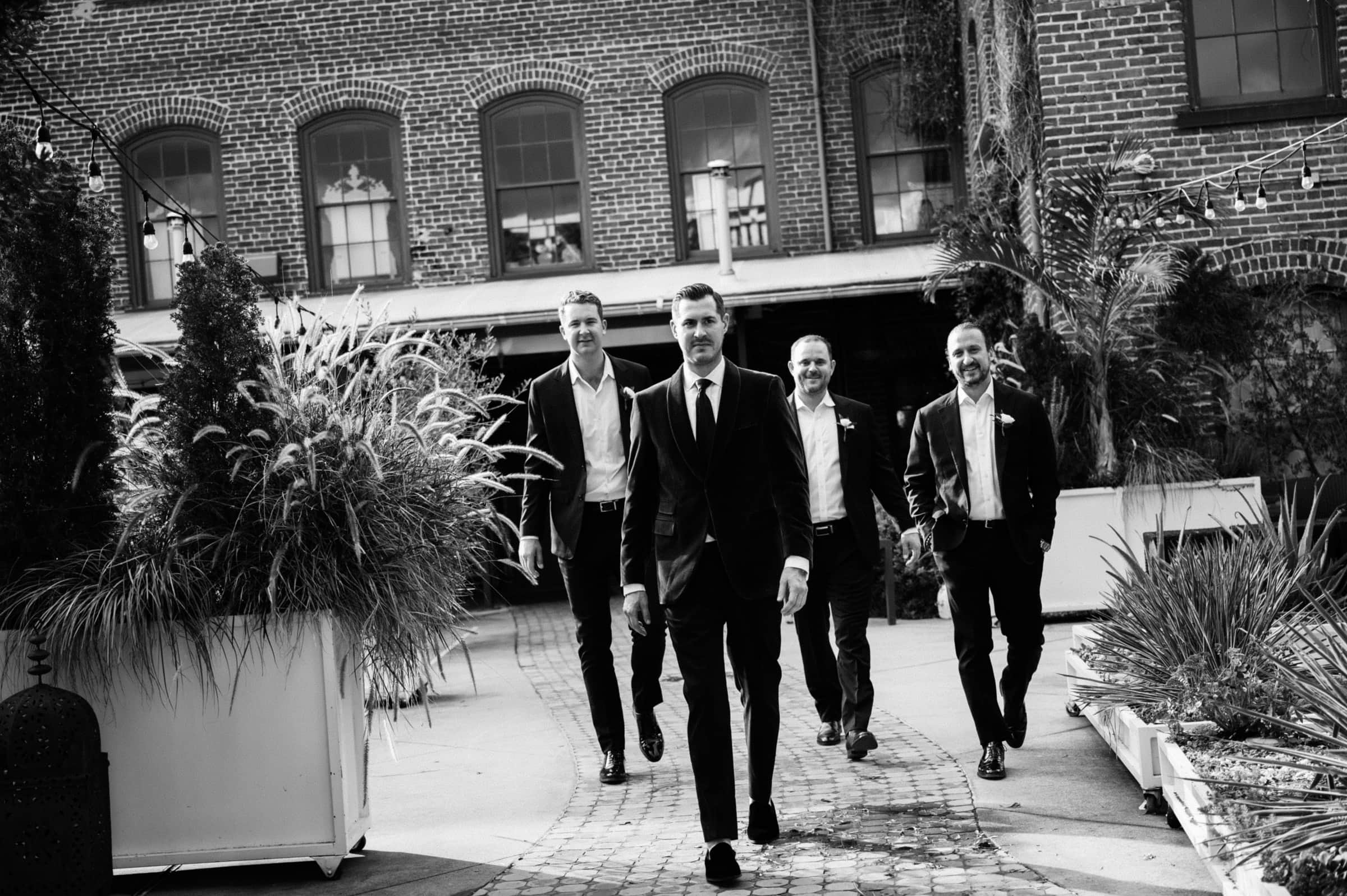 Four men in formal attire walk confidently in a courtyard at Melrose Knitting Mill, surrounded by brick buildings and lush greenery. The black-and-white scene highlights the textures of their outfits, capturing the timeless elegance of a wedding day moment.