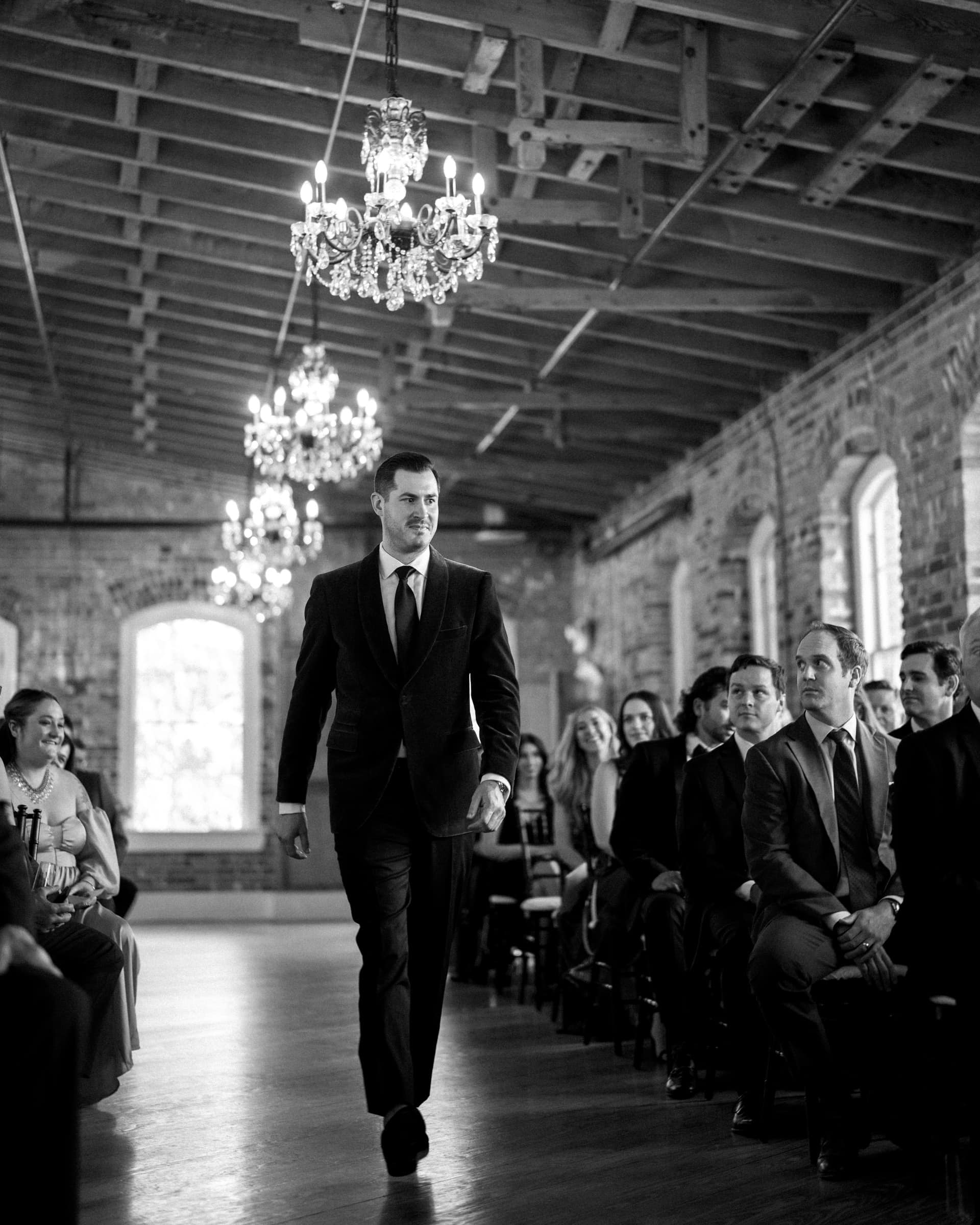 A man in a suit walks down the aisle at Melrose Knitting Mill, a rustic venue with chandeliers overhead. Formally dressed guests watch him in anticipation amidst brick walls, large windows, and a wooden ceiling.