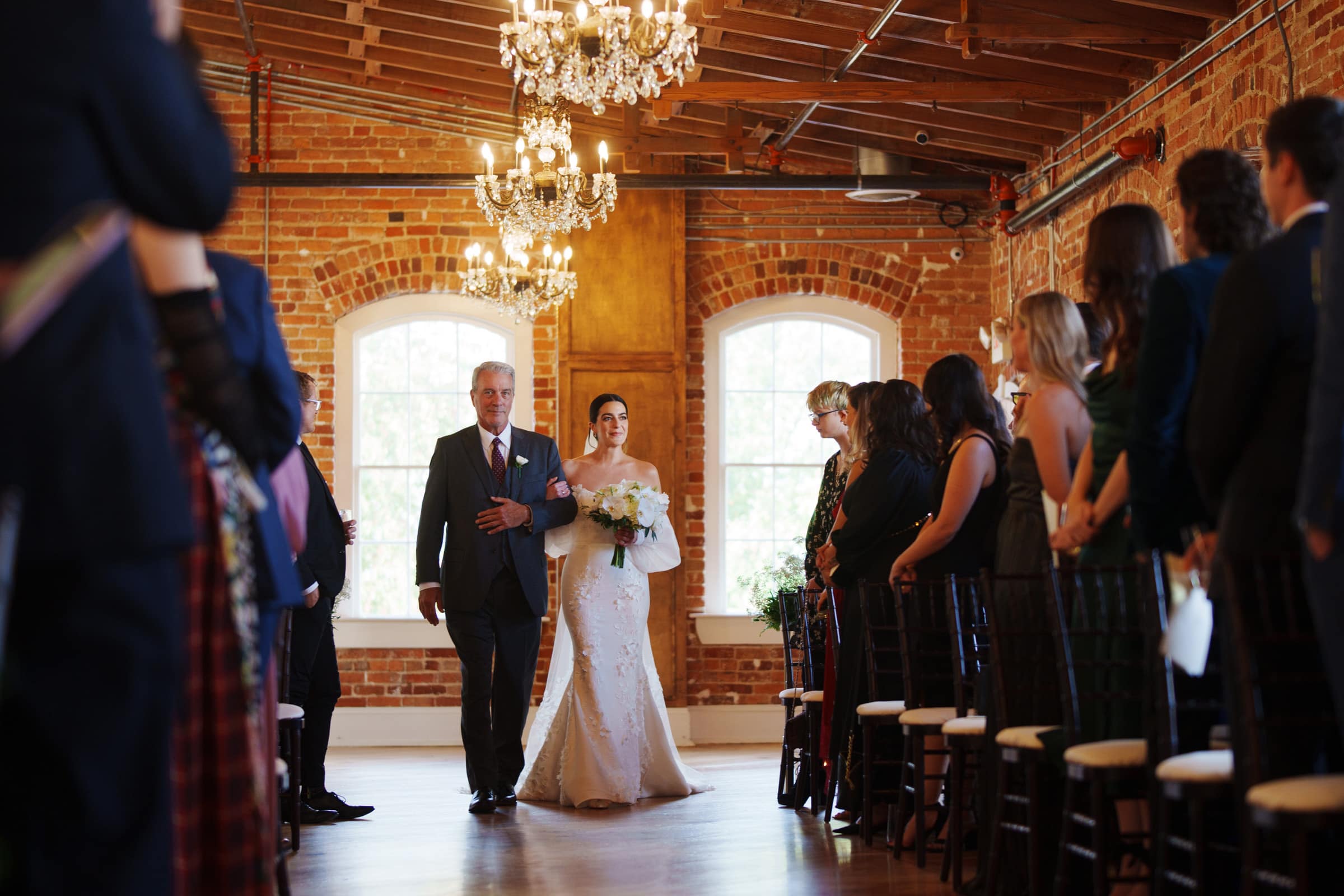 A bride in a white dress walks down the aisle with a man in a suit, possibly her father, at the charming Melrose Knitting Mill wedding venue. They are surrounded by brick walls and chandeliers, while guests seated on either side watch the procession.