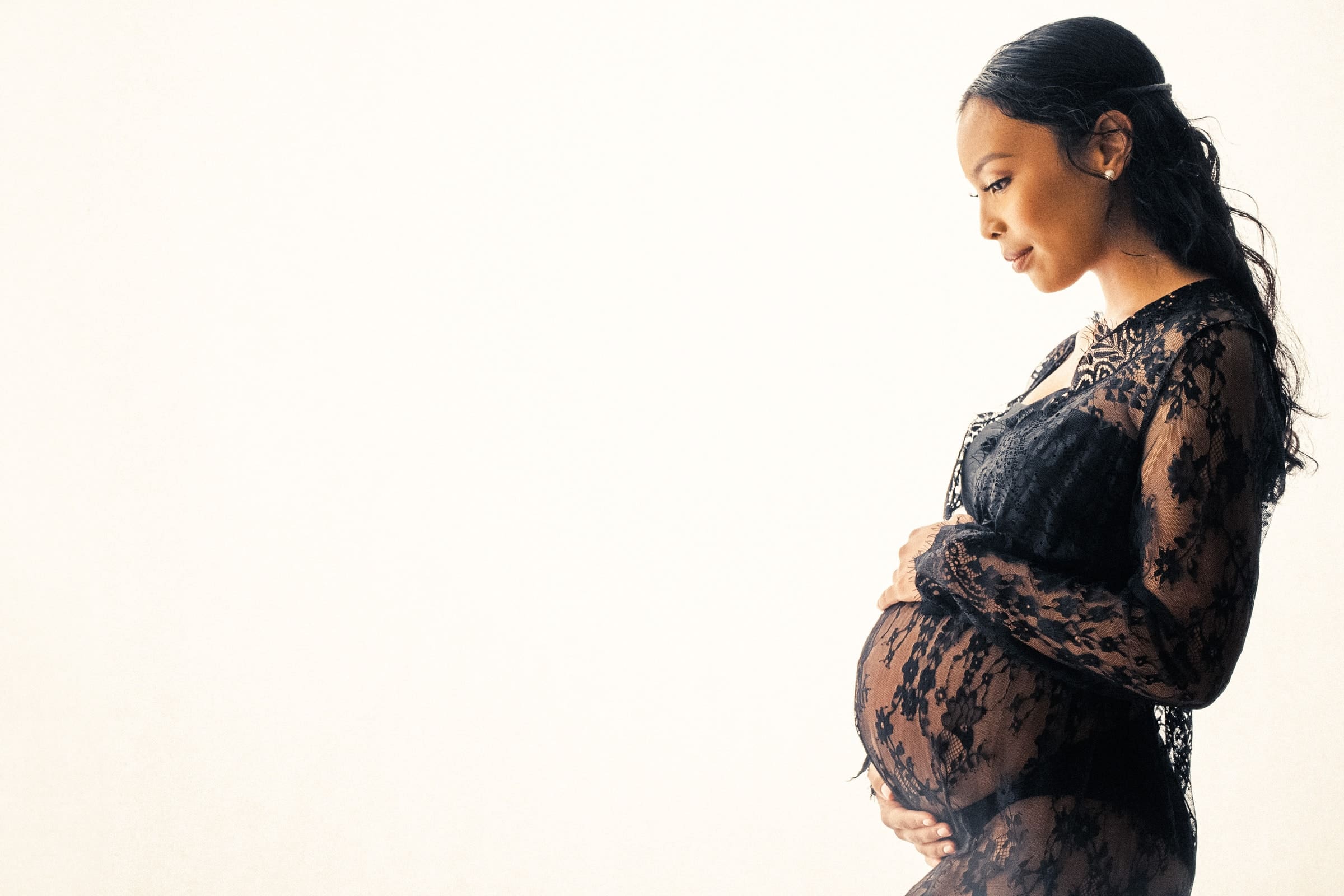 A pregnant woman in a black lace dress gently cradles her belly, standing against a light background. She appears serene and thoughtful, gazing downward. This Raleigh maternity photography captures a tender and intimate moment.